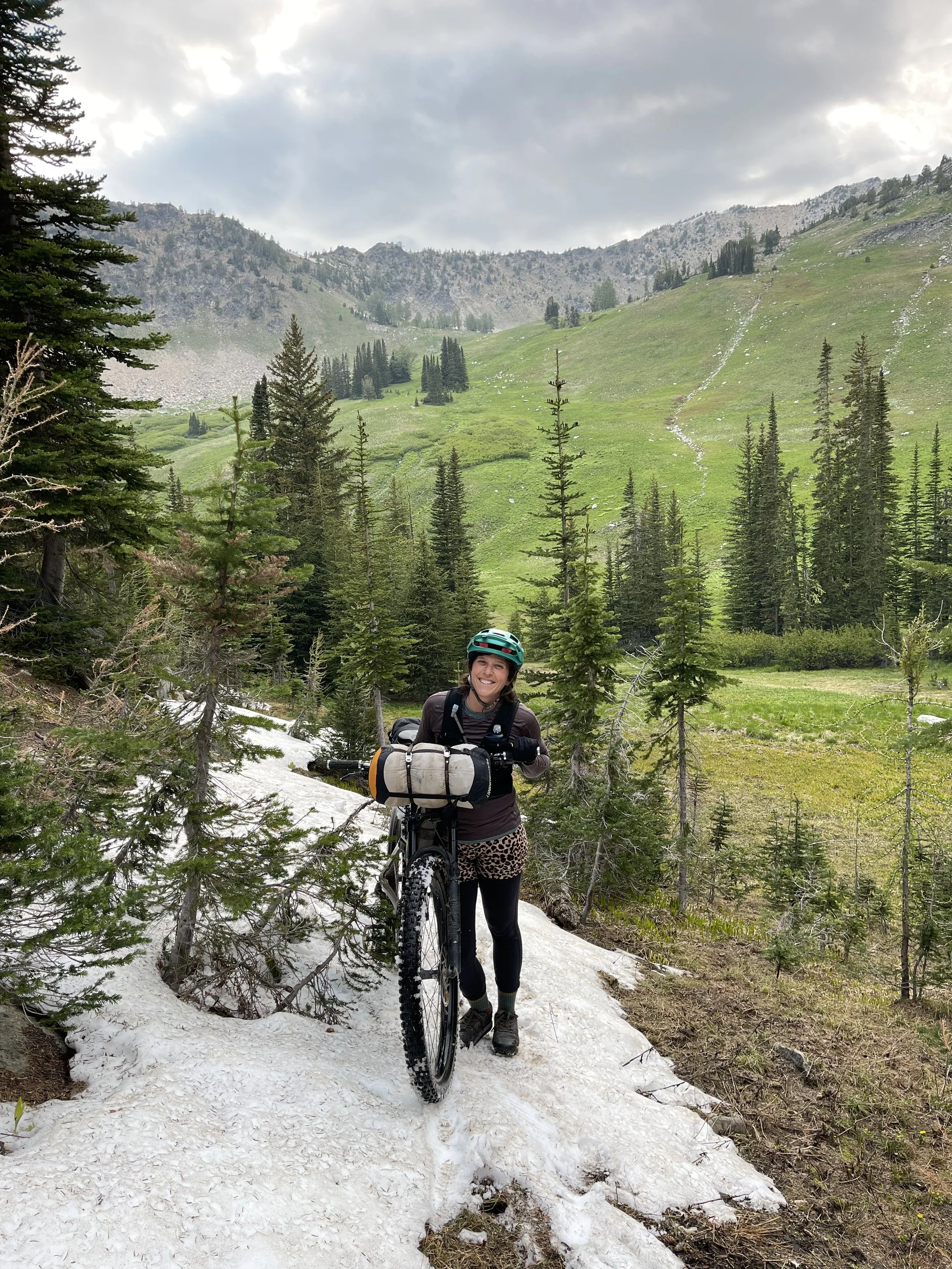A woman wearing a teal bike helmet, dark jacket, and leopard print pants smiles while standing with her mountain bike on a snowy trail in a mountainous forest area. Green hills, tall pine trees, and gray, clouded sky are in the background.
