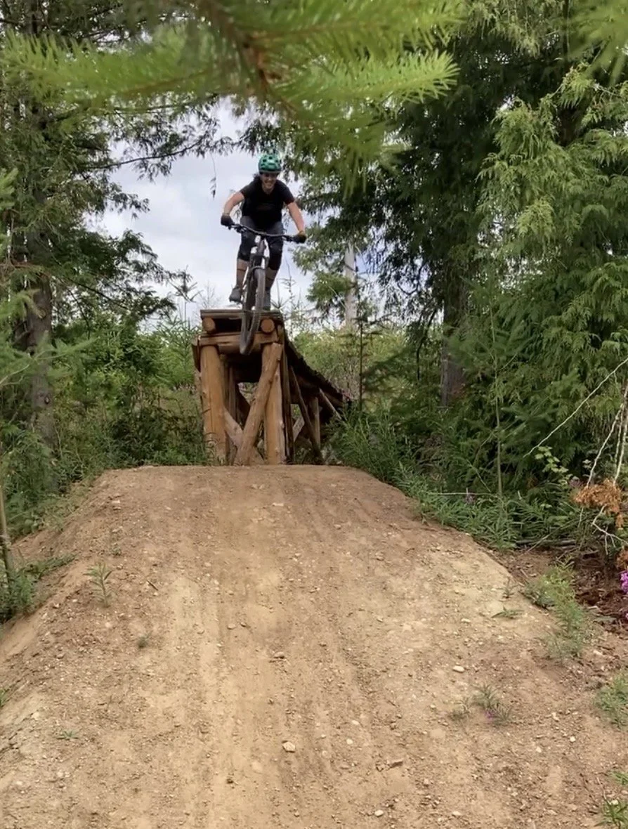 Person riding a mountain bike down a dirt jump ramp in a forested area.