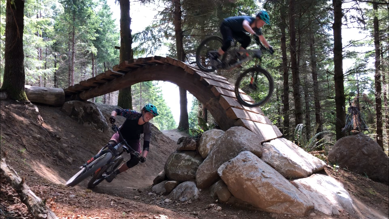 Two mountain bikers riding on a forest trail, one riding under a wooden bridge and the other leaning into a turn on the dirt path surrounded by trees and large rocks.
