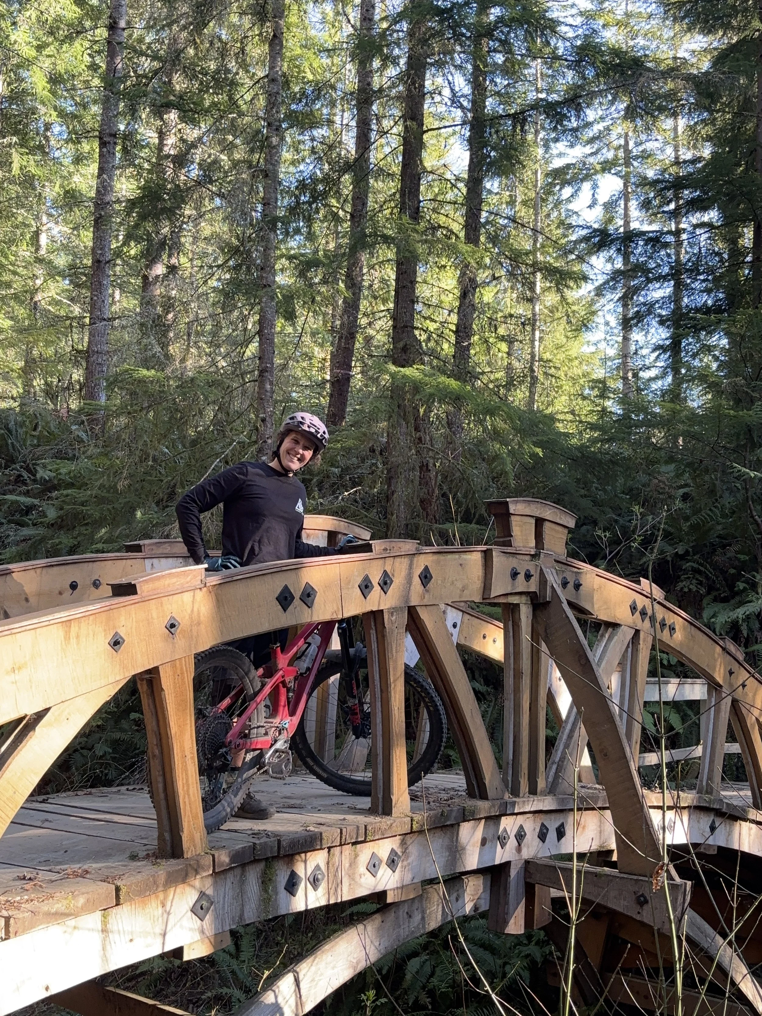 A person wearing a helmet and black clothing standing on a wooden bridge with a bicycle attached underneath, surrounded by tall green trees in a forest setting.