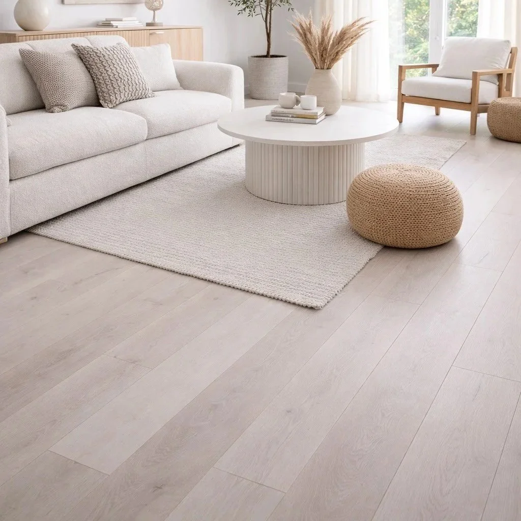 A bright, minimalist living room with a light-colored sofa, a round white coffee table, a woven pouf, a beige armchair, and decor including a large potted plant, vases with dried plants, and books, with natural light coming through sheer curtains.