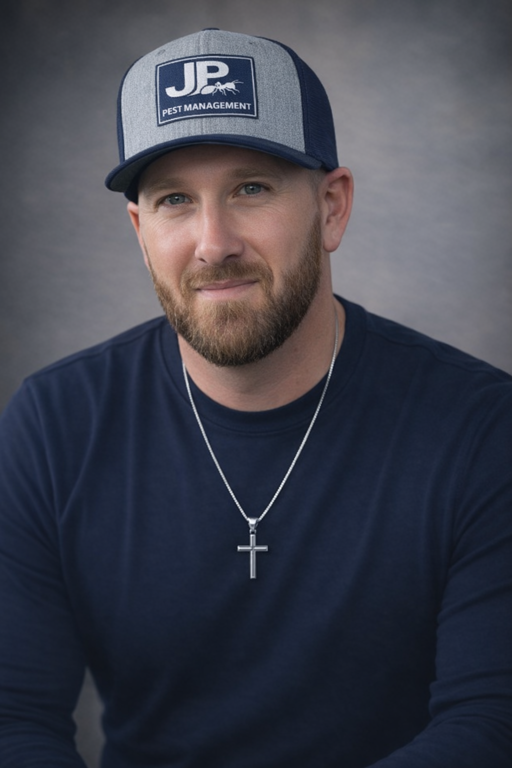 A man with a beard and blue eyes wearing a navy blue shirt, a silver cross necklace, and a gray and navy baseball cap with a logo that reads 'JP Pest Management'.