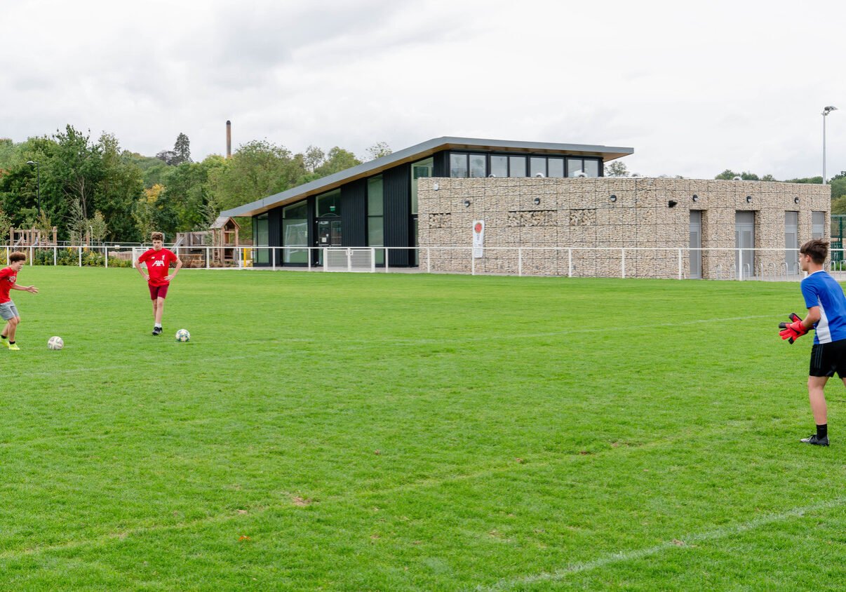 Three boys playing soccer on a green field near a modern building, with trees and a cloudy sky in the background.