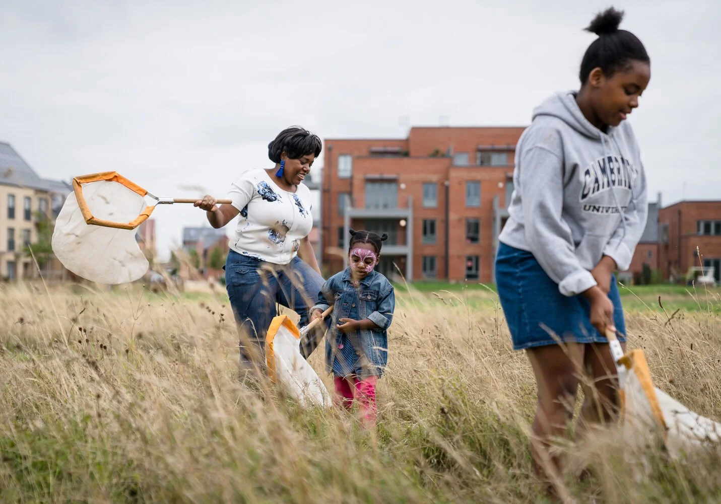 Four people, including an adult woman and two young girls, are collecting trash in a grassy field with nets. The woman is smiling, and the girls are focused on their task. There are residential buildings in the background.