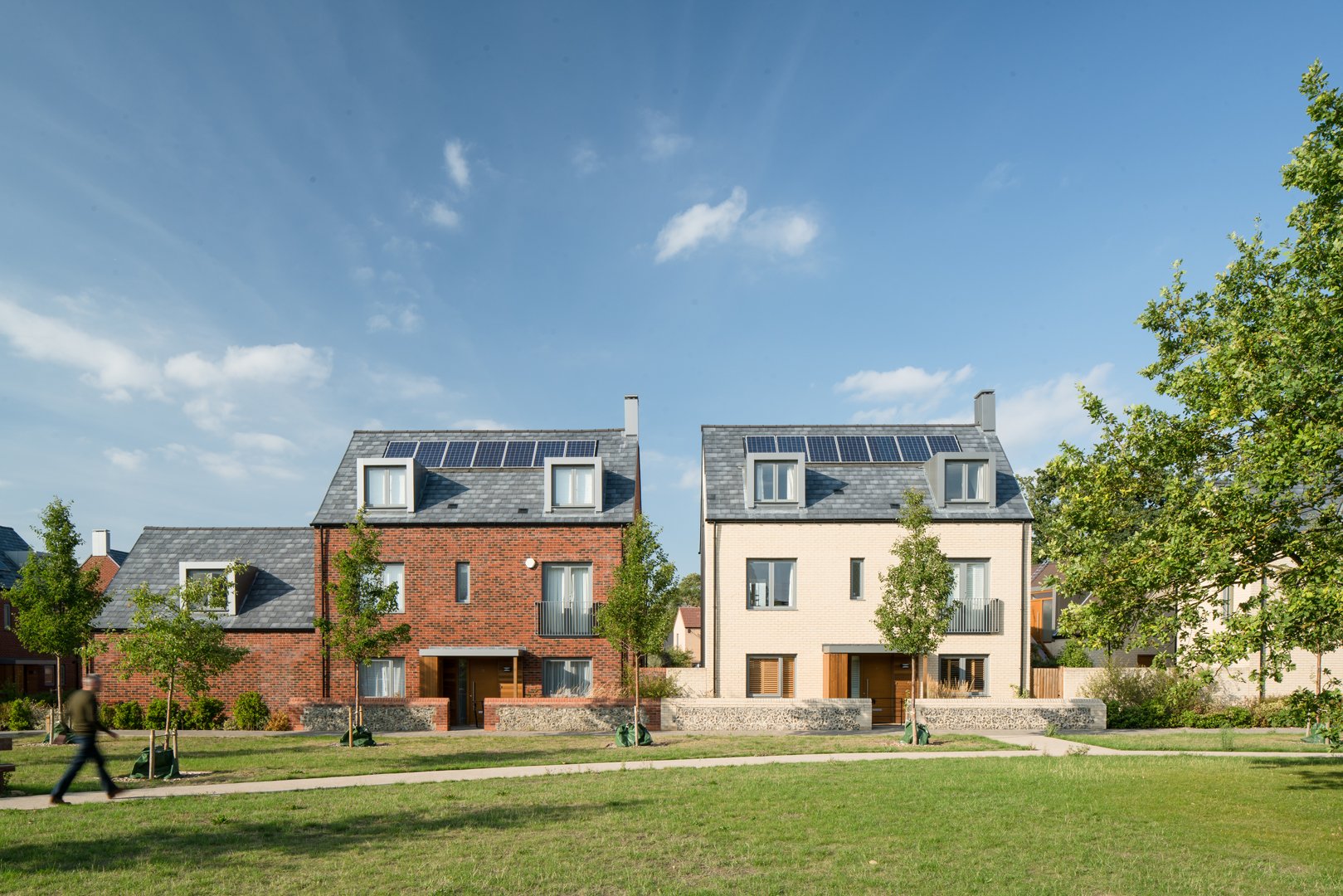 Two modern houses with solar panels on rooftops, situated in a grassy area with trees and walking path under a blue sky.