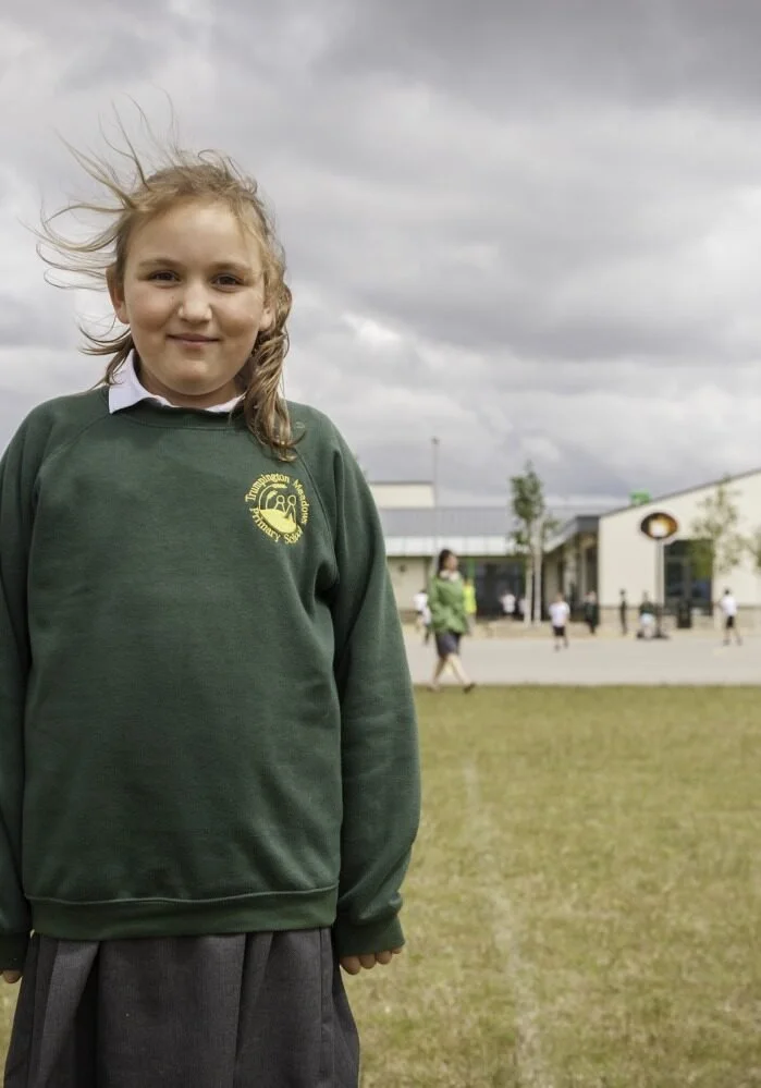 A young girl with blonde hair standing outside on a cloudy day, wearing a green school sweatshirt with a logo, and a gray skirt.