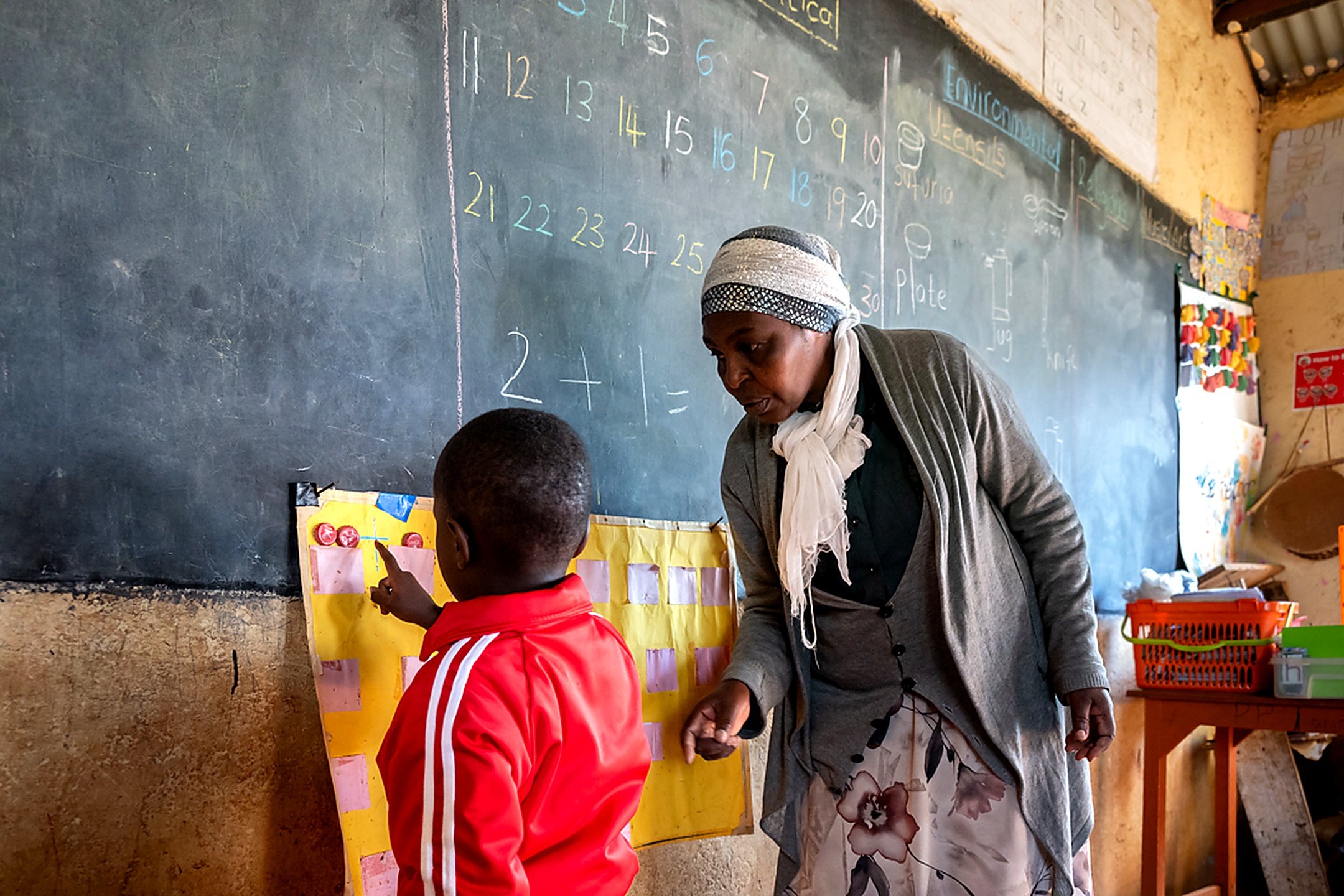A teacher or caregiver talking to a young student in a classroom, with a chalkboard displaying numbers and writing in the background. The student is wearing a red jacket and pointing at a chart on the wall.