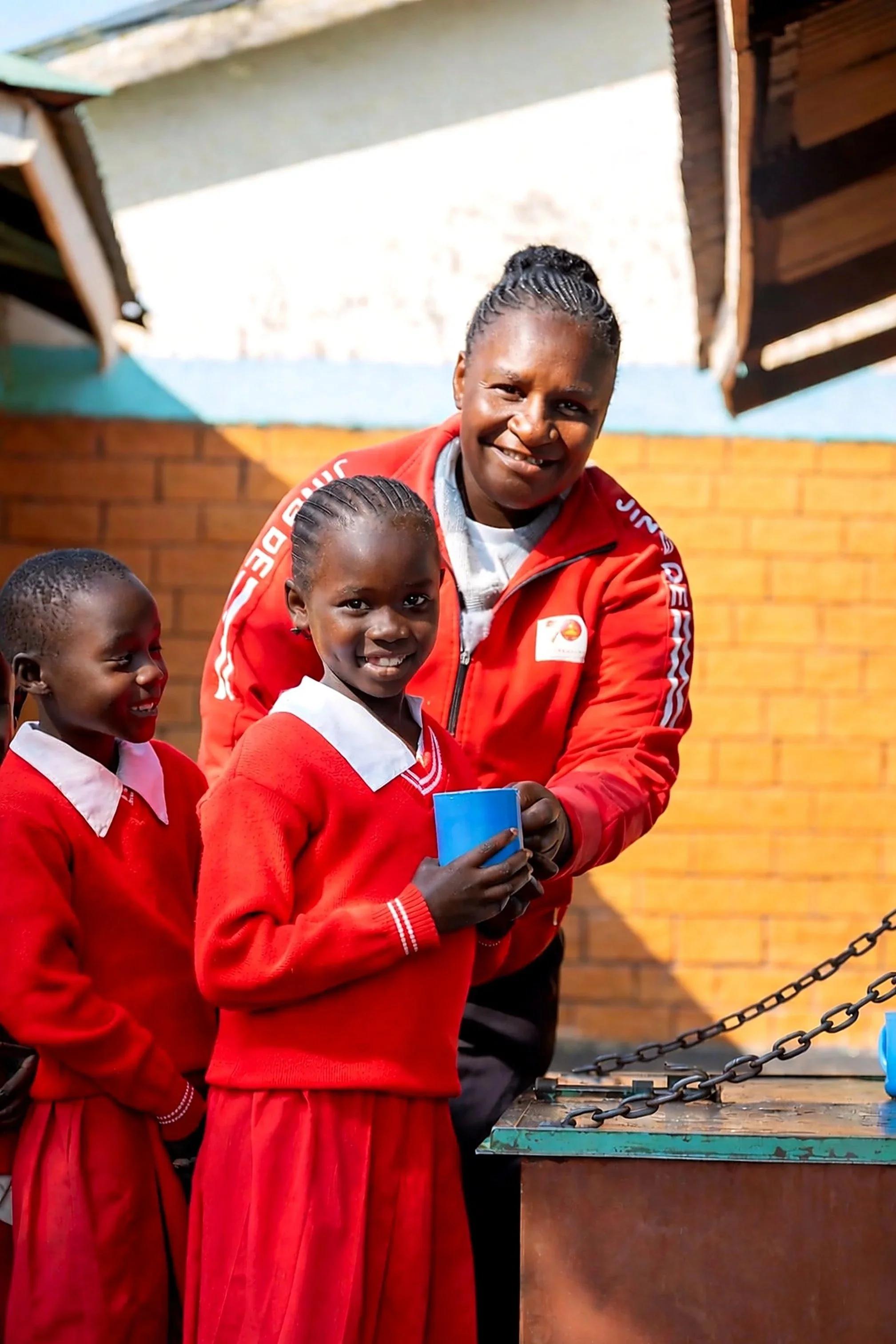 A woman and two girls wearing red school uniforms smiling, standing outdoors behind a table with a chain link barrier, against a brick wall background.
