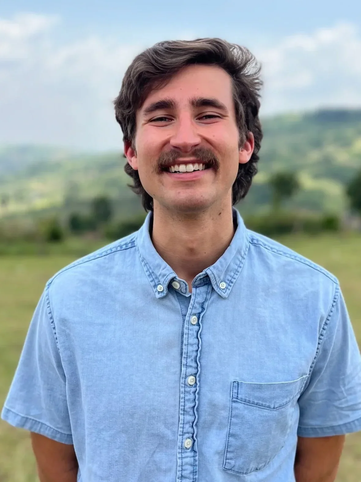 Smiling mustached man in a light blue button-down shirt standing outdoors with a blurred green landscape background