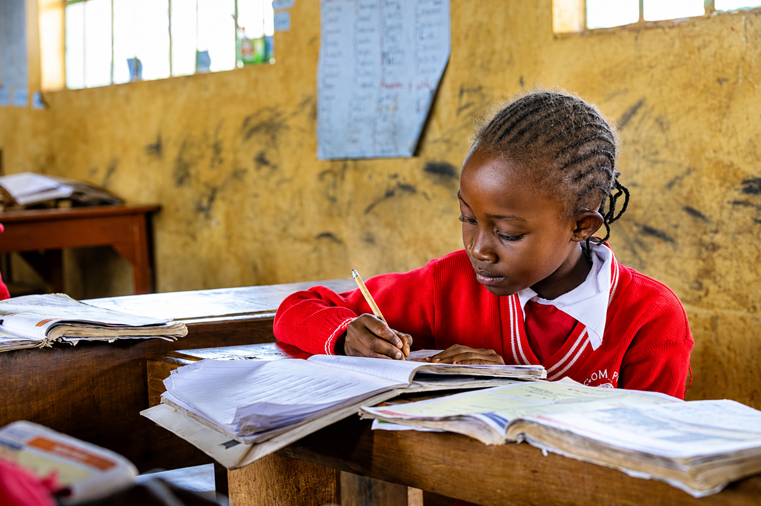 A young girl in a red school uniform studying at her desk in a classroom with a yellow wall and posters.