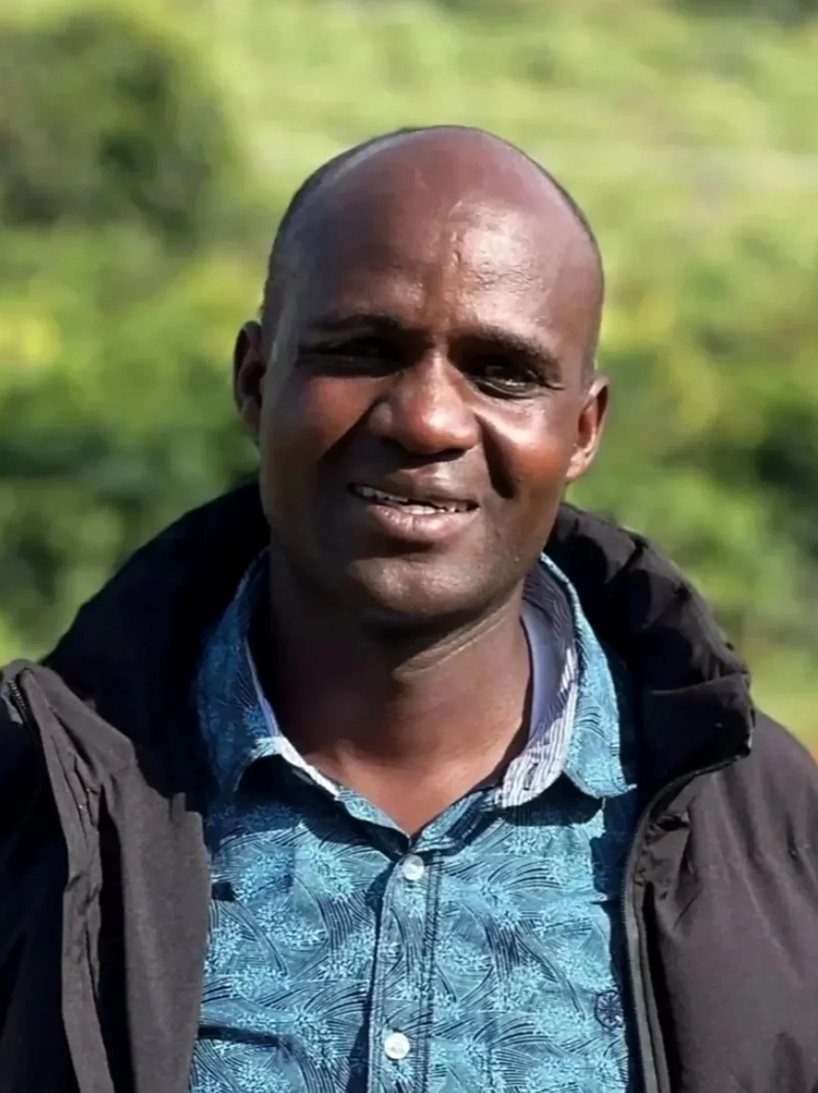 Close-up of a smiling man with a shaved head wearing a patterned blue shirt and a dark jacket, outdoors with green foliage in the background.