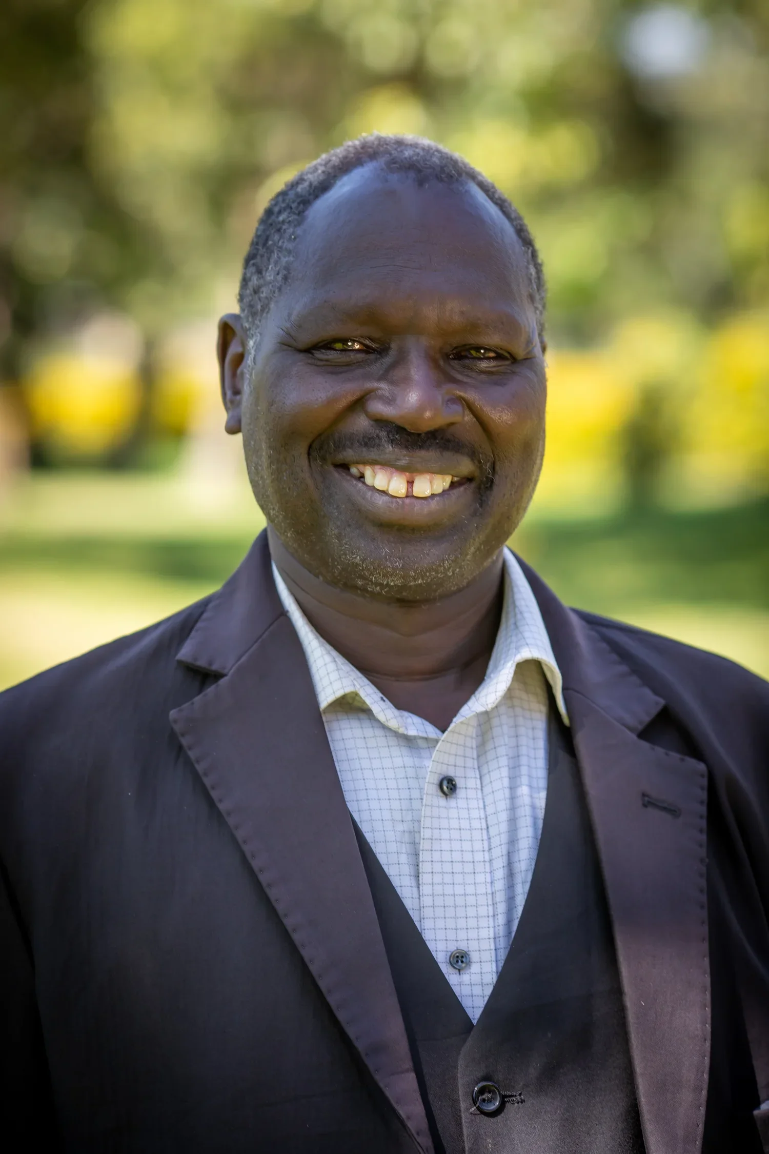 portrait of a smiling man in a suit outdoors, with blurred green trees in the background.