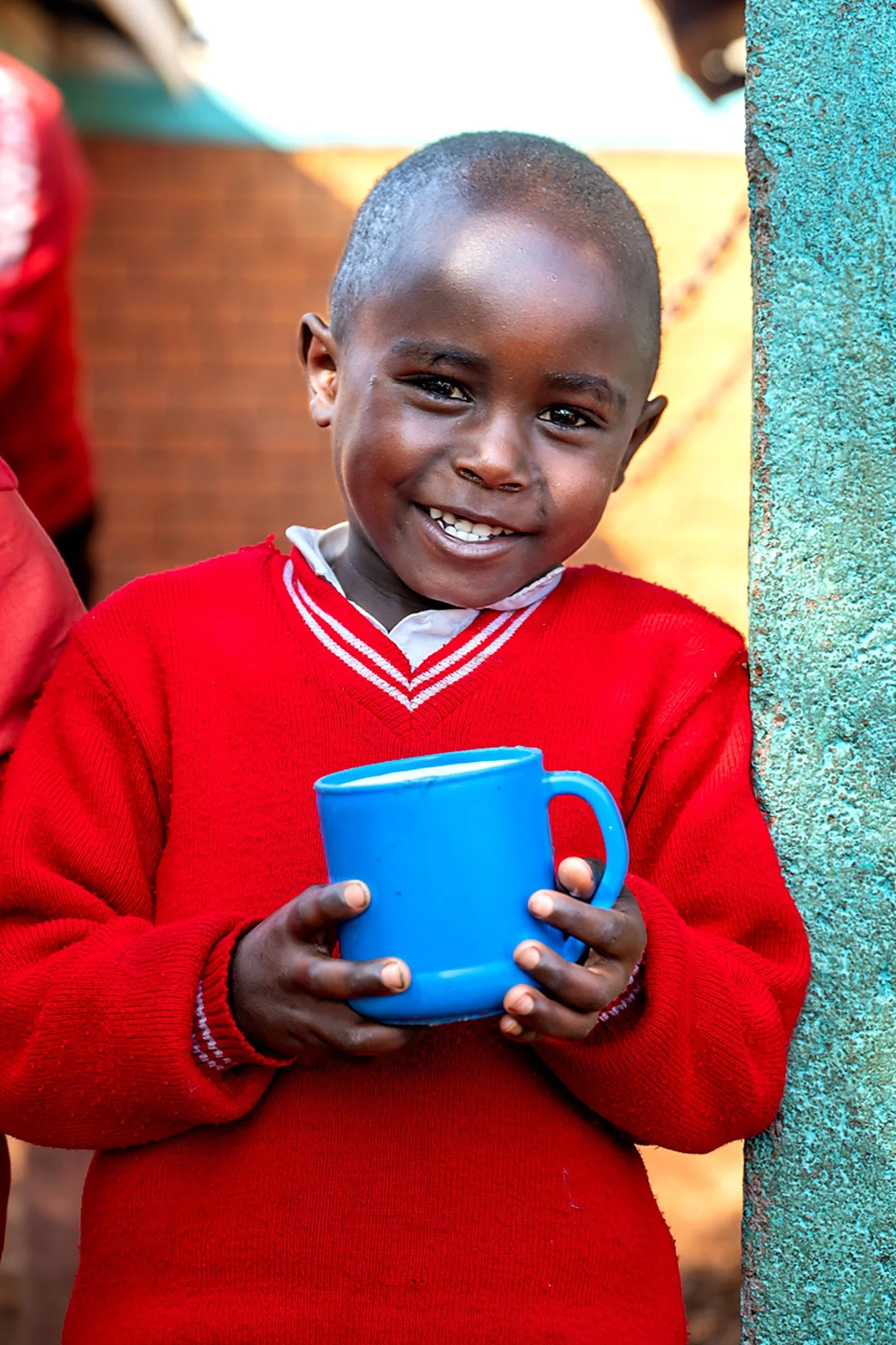 A young boy smiling, wearing a red sweater and holding a blue mug, standing outdoors next to a textured green wall.