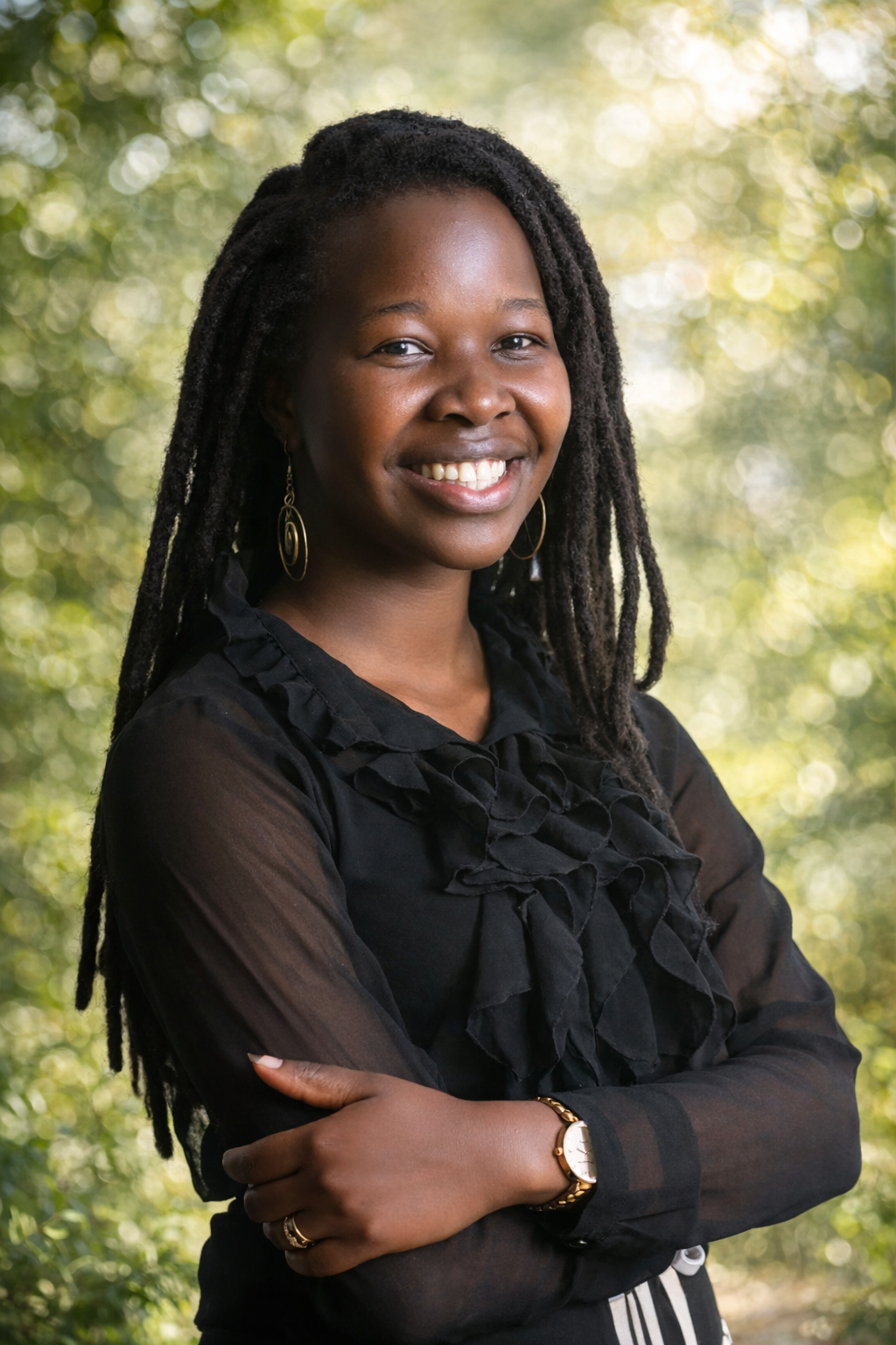 A woman with long dreadlocks, wearing a black blouse with ruffled details, gold hoop earrings, a gold watch, and rings, stands outdoors with trees and sunlight in the background, smiling with her arms crossed.
