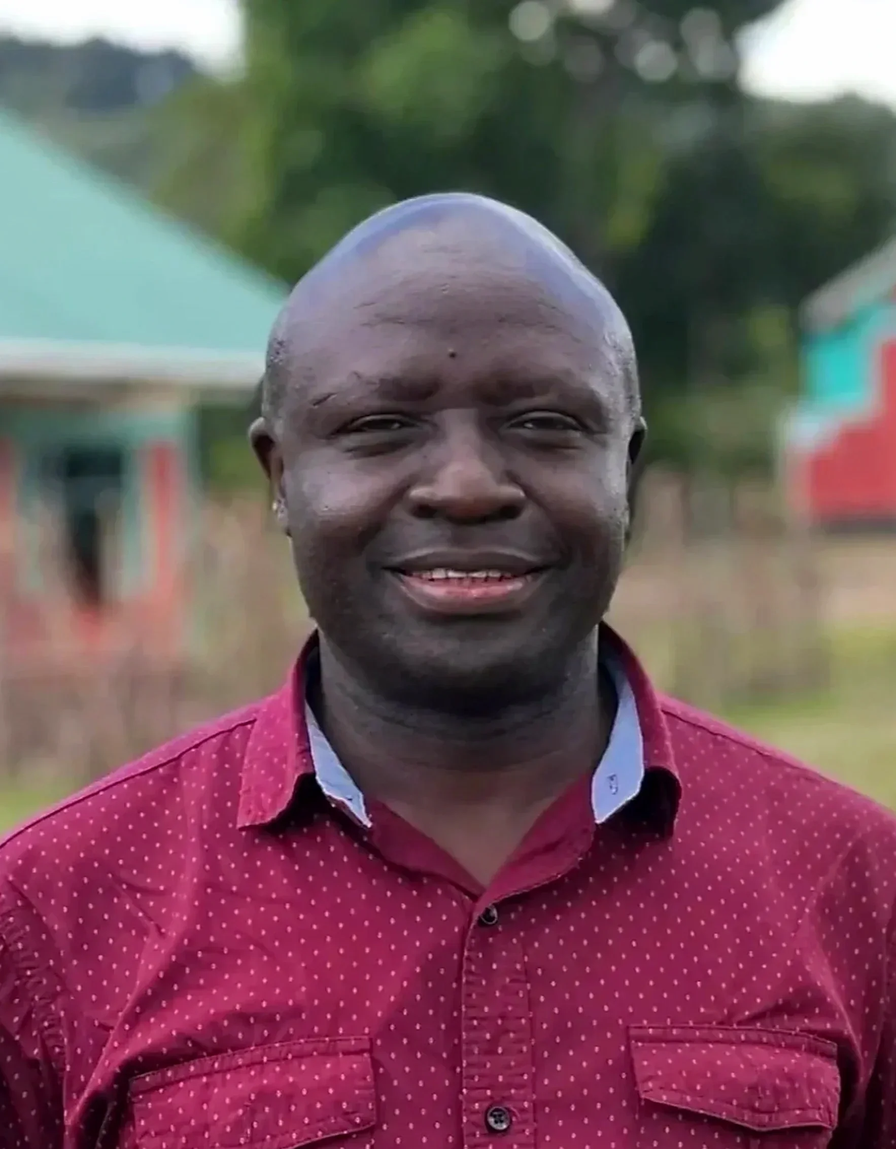 Close-up of a smiling man with a shaved head, wearing a red shirt with small white polka dots, outdoors with green trees and blurry buildings in the background.