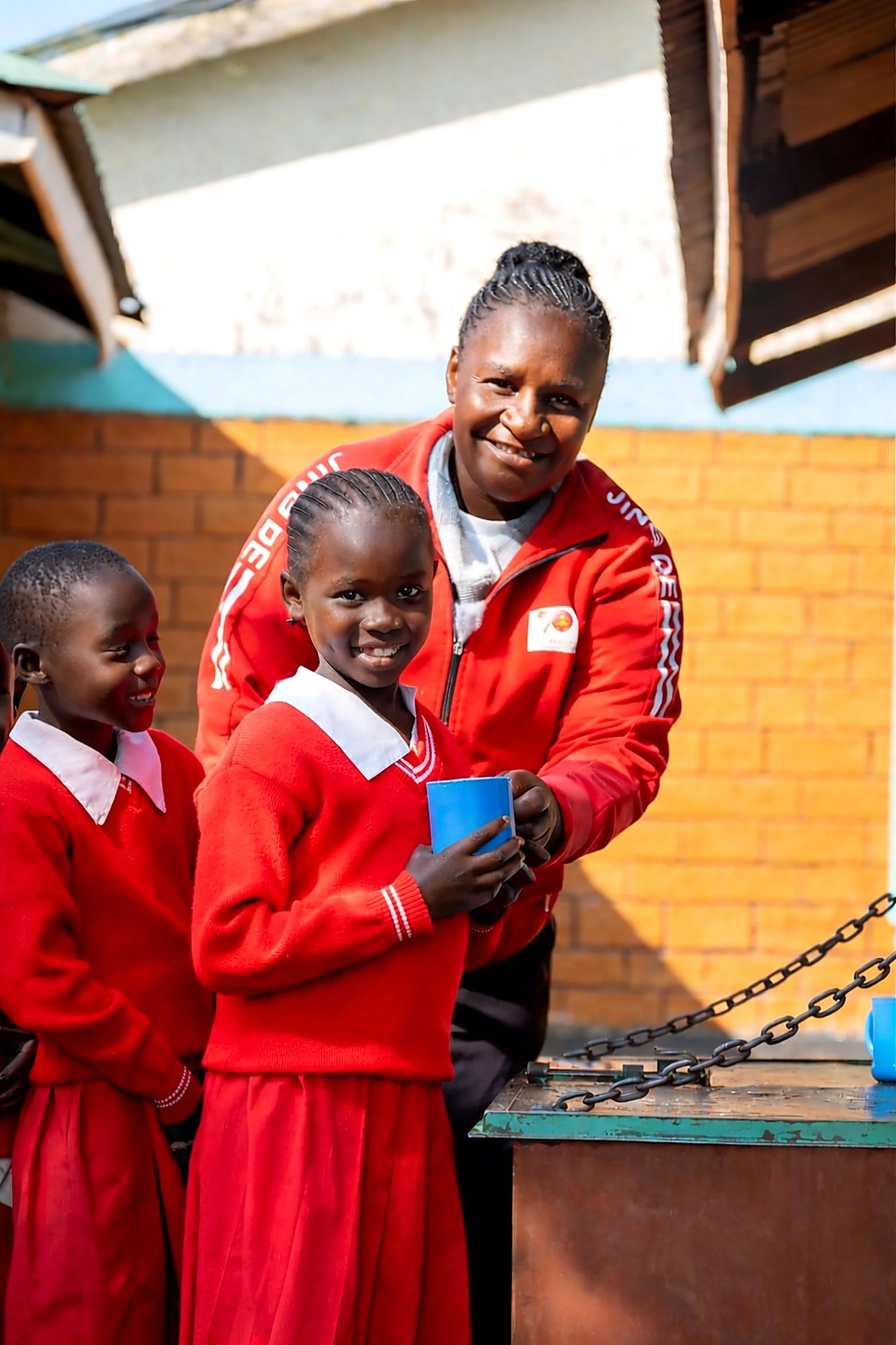Three school children wearing red uniforms and an adult woman, smiling at the camera, standing outdoors with a brick wall and wooden shelter in the background.