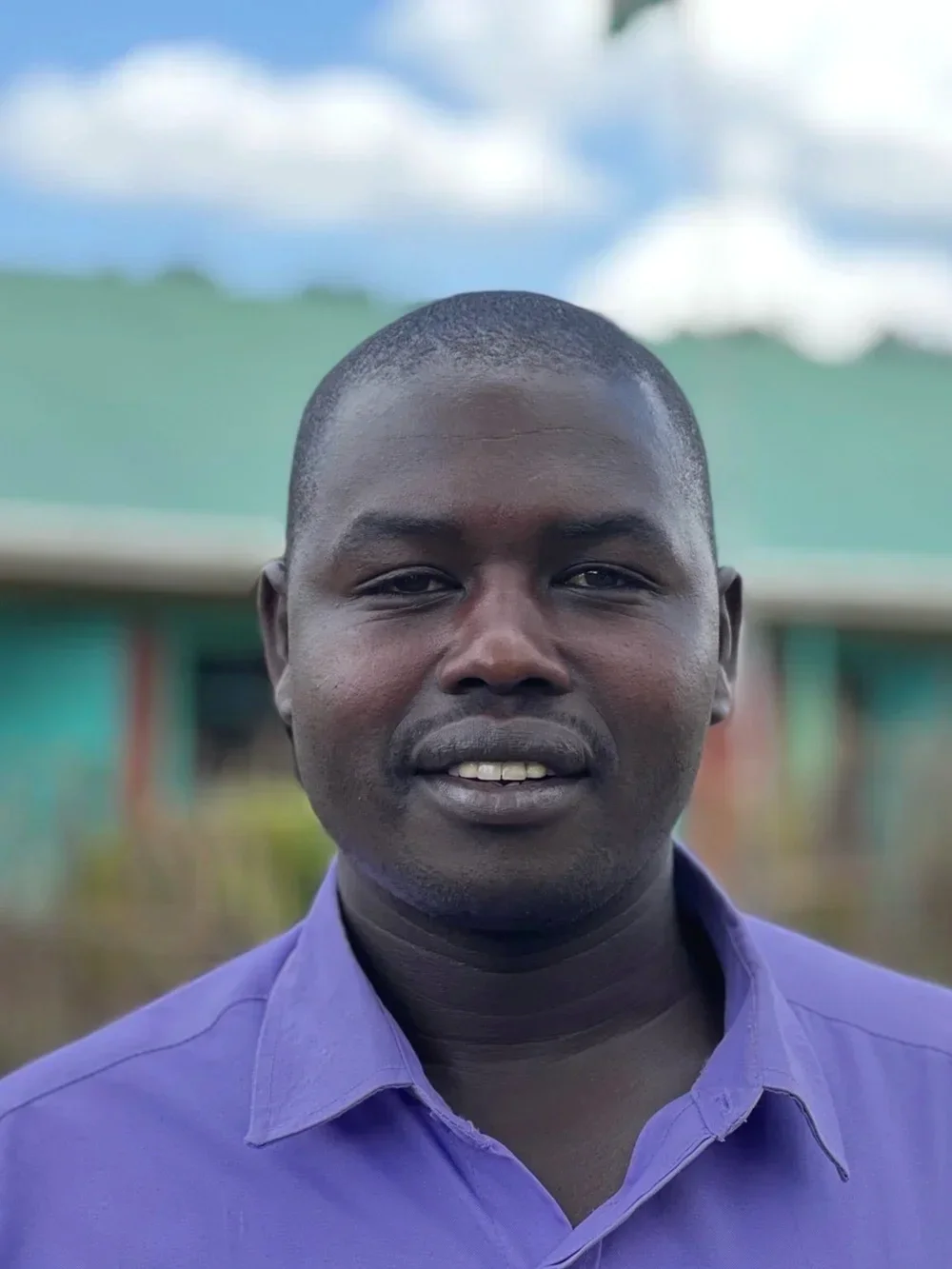 A man with dark skin wearing a purple shirt, standing outdoors with a blurred green and blue background, smiling at the camera.
