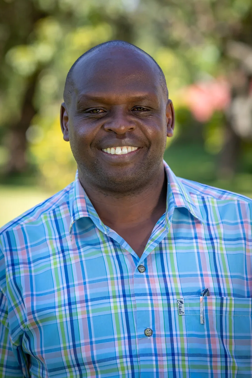A smiling Black man with short hair wearing a colorful plaid button-up shirt, standing outdoors with blurred greenery in the background.