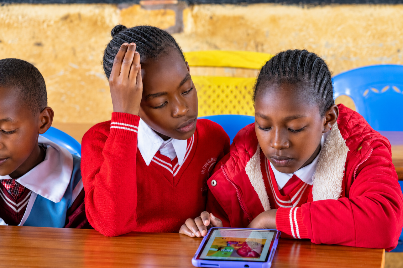Three schoolchildren in red uniforms sit at a classroom desk, looking at a tablet.