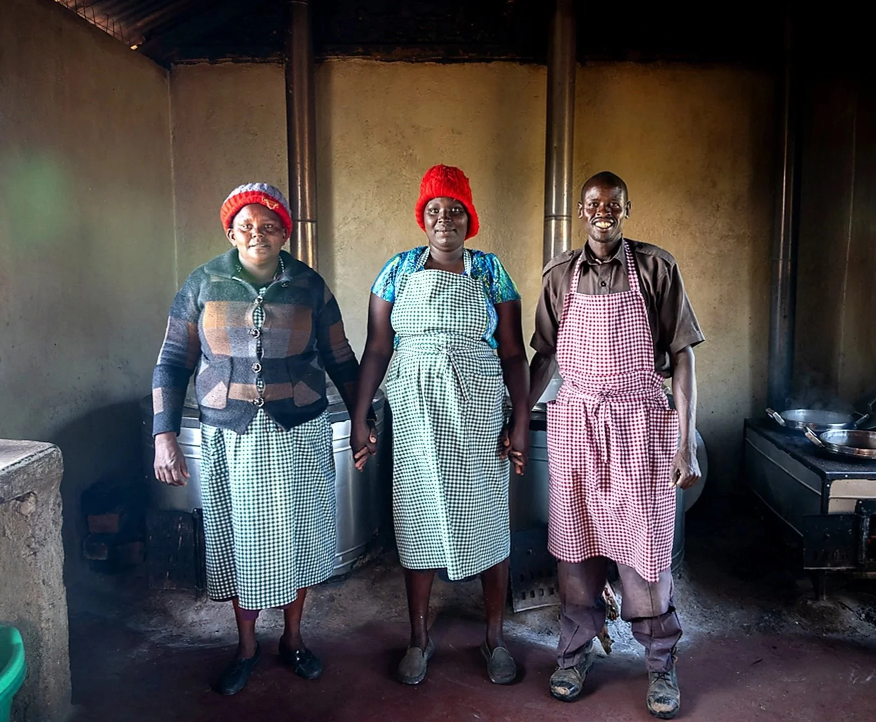 Three women and one man standing in a kitchen, wearing aprons and warm clothing, with cooking pots visible in the background.