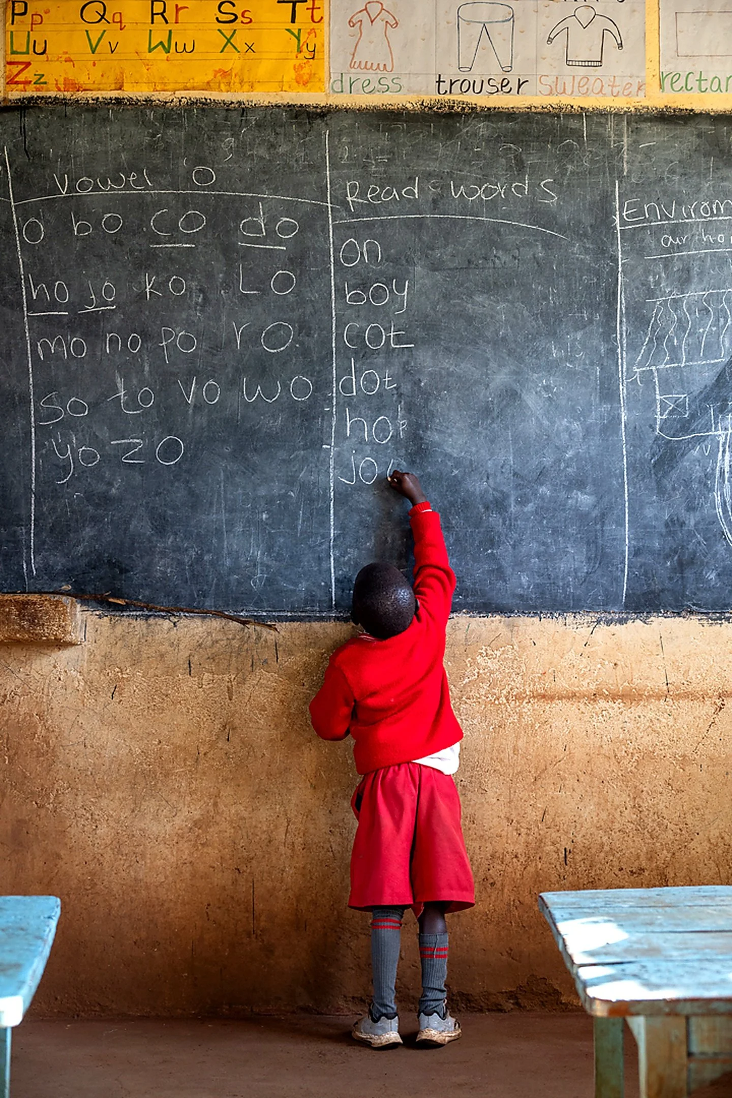 A young student in a red school uniform with gray knee-high socks and sneakers, standing at a blackboard and writing with chalk in a classroom. The classroom has a yellow wall with educational posters and a brown wall below.