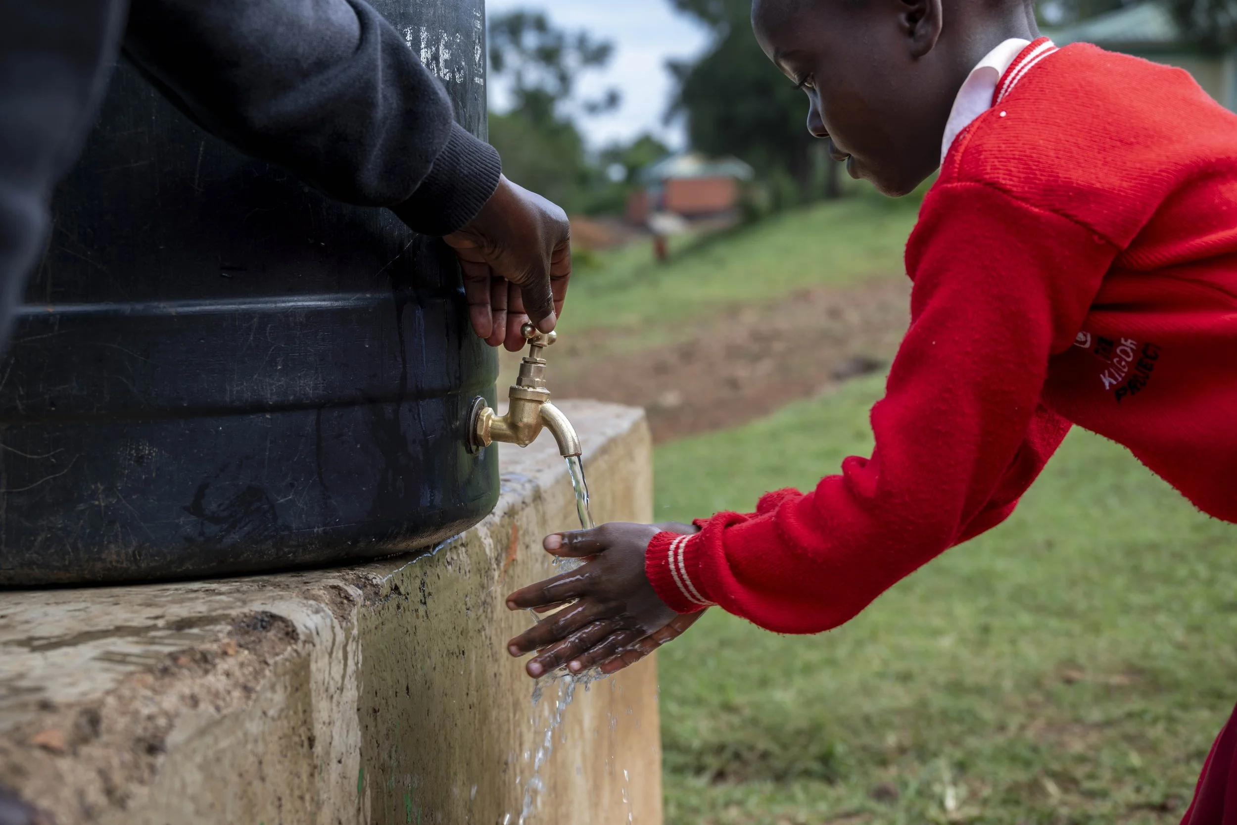 A young boy in a red sweater washing his hands under running water from an outdoor faucet near a water tank, with a green landscape in the background.