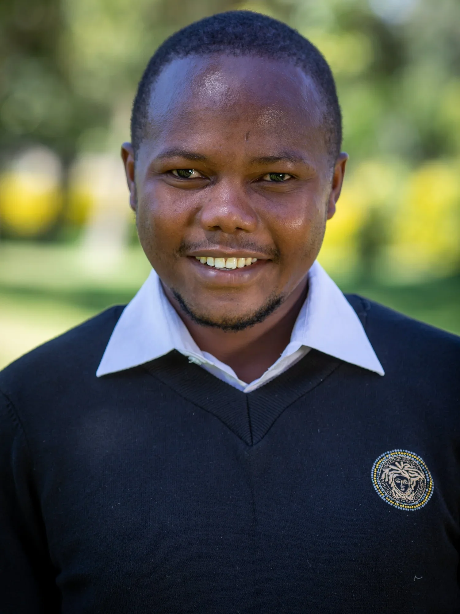 Close-up of a smiling young man with short hair wearing a white collared shirt and a black sweater with an embroidered emblem on the chest, outdoors with blurred green foliage in the background.