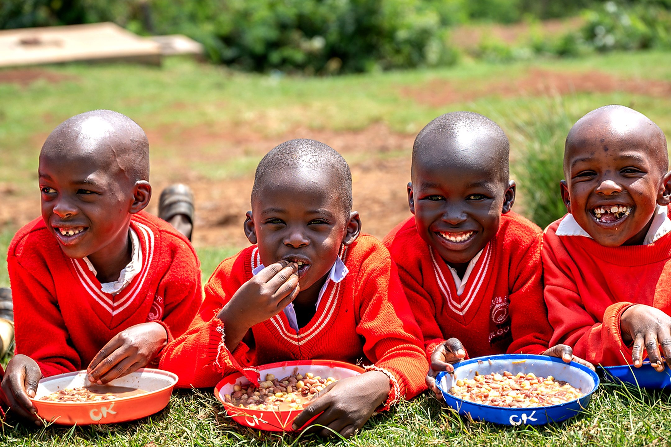 Four young children sitting outdoors on grass, smiling and eating from bowls of food, wearing red sweaters with white shirts underneath.