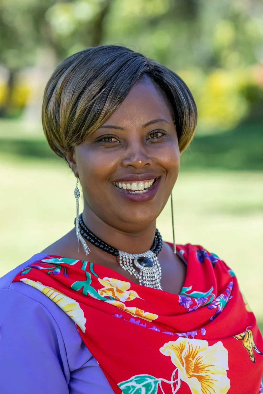 A woman smiling outdoors with a blurred green background, wearing a red floral scarf, purple top, and jewelry.