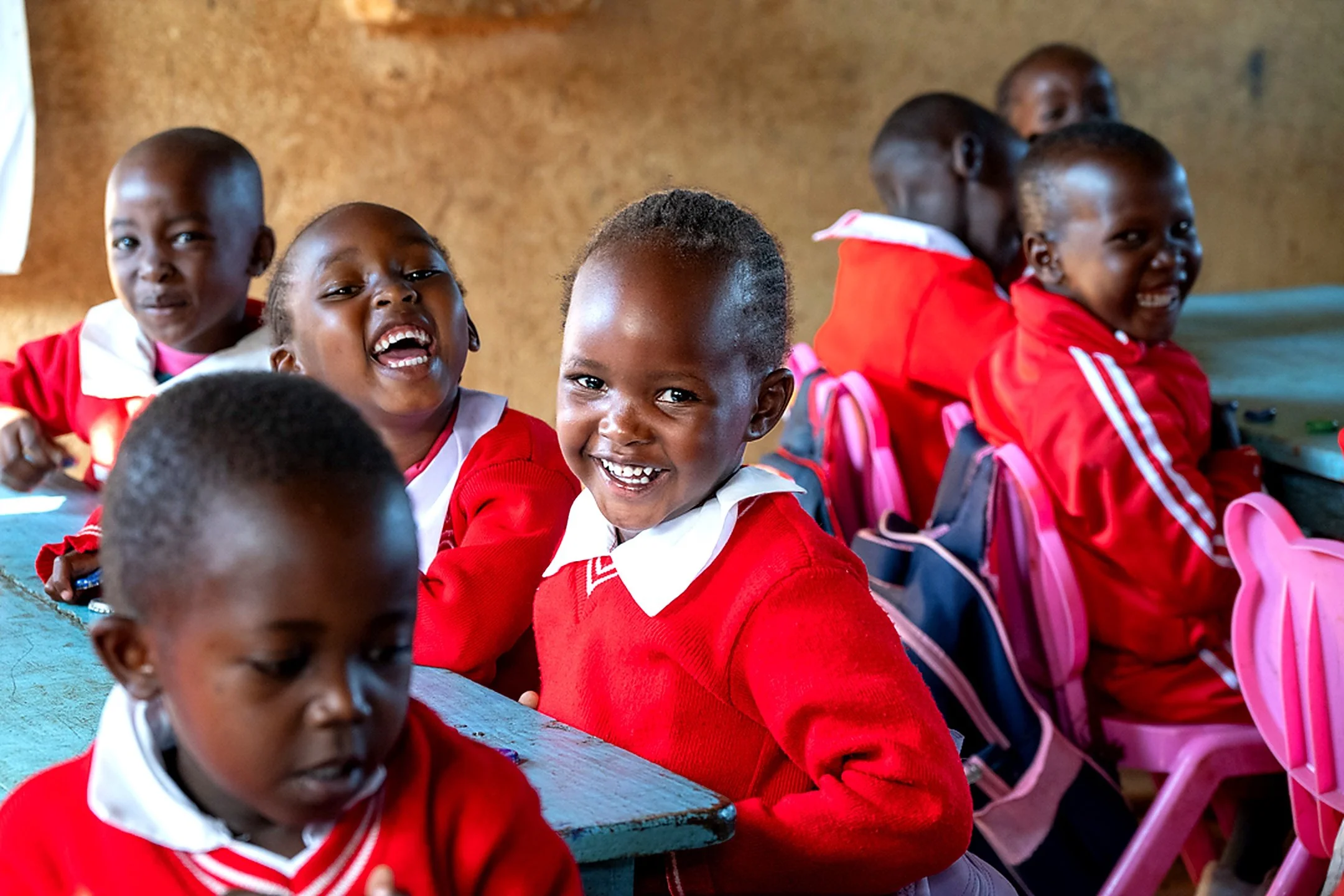 A group of young African children dressed in red and white school uniforms sitting in a classroom, smiling and laughing.