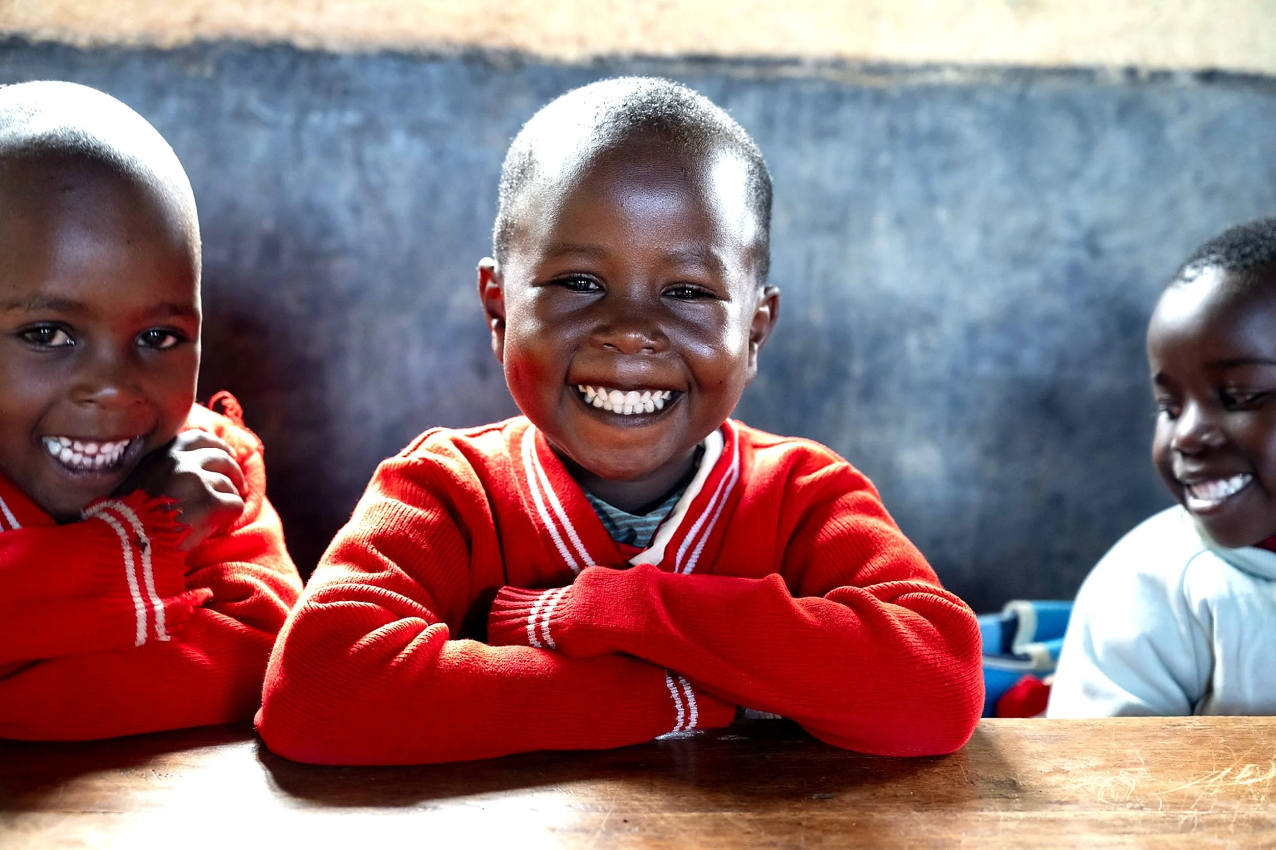 Smiling children sitting at a table, with a focus on a child wearing a red sweater in the center.