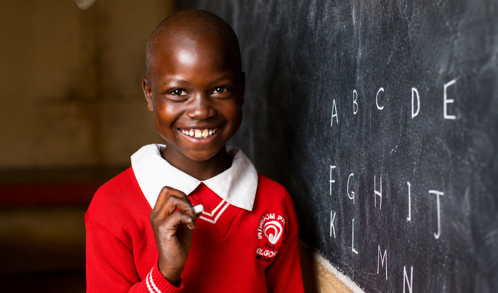 A smiling young girl with short hair in a red school uniform standing next to a blackboard with the alphabet written on it.