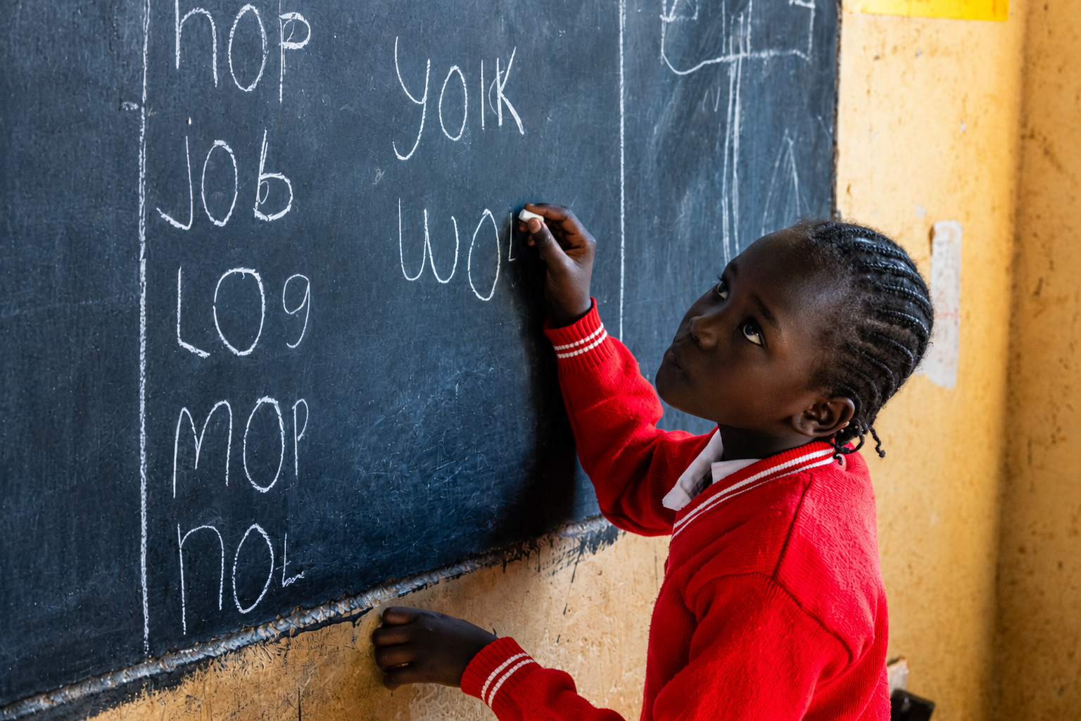 A young girl writing on a school blackboard with white chalk, with words like "top," "job," and "mop" listed vertically, and her focusing on writing the word "work."