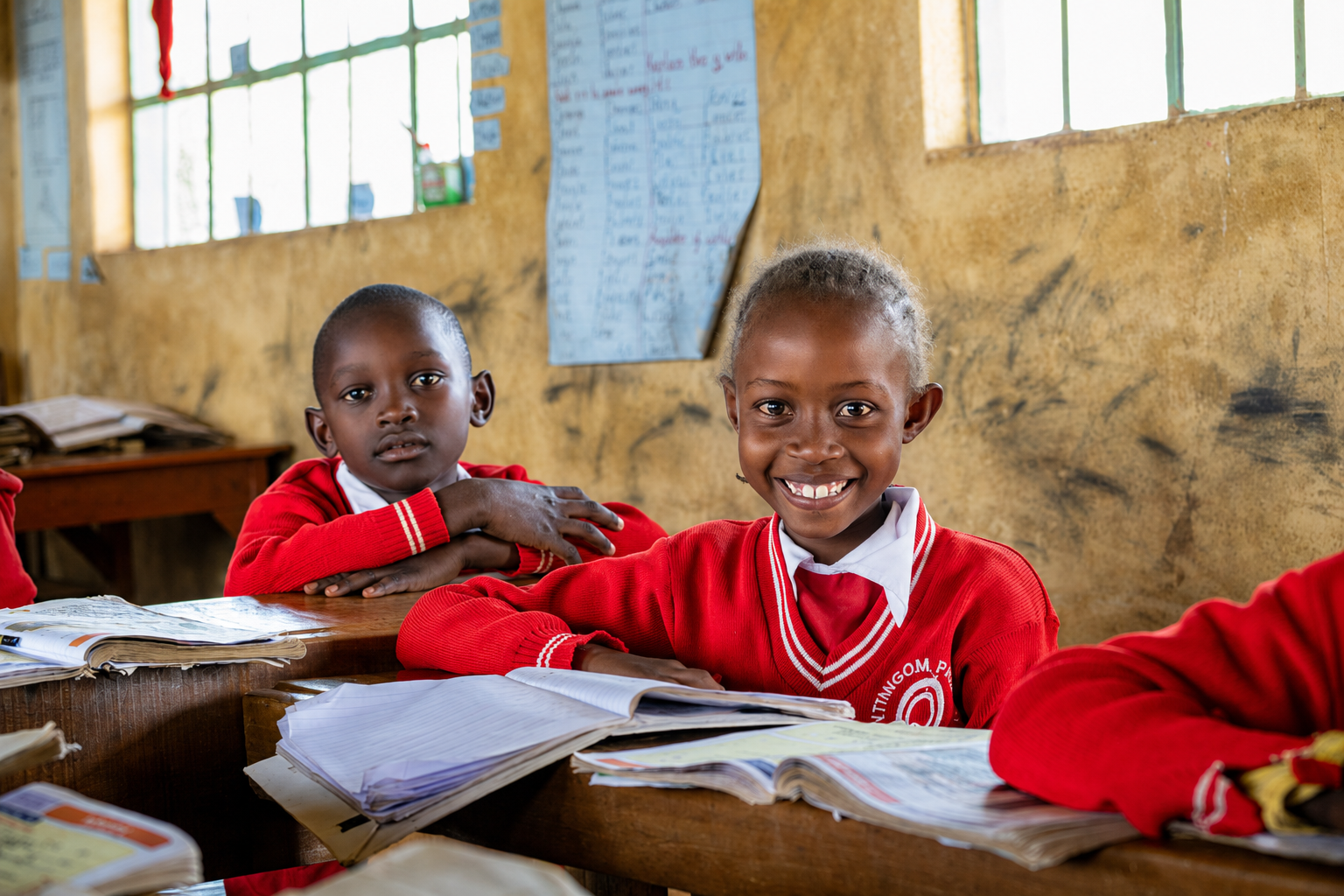 Two young students in red school uniforms sitting at desks inside a classroom with yellow walls, books and notebooks in front of them, and a large paper chart with writing on the wall behind.