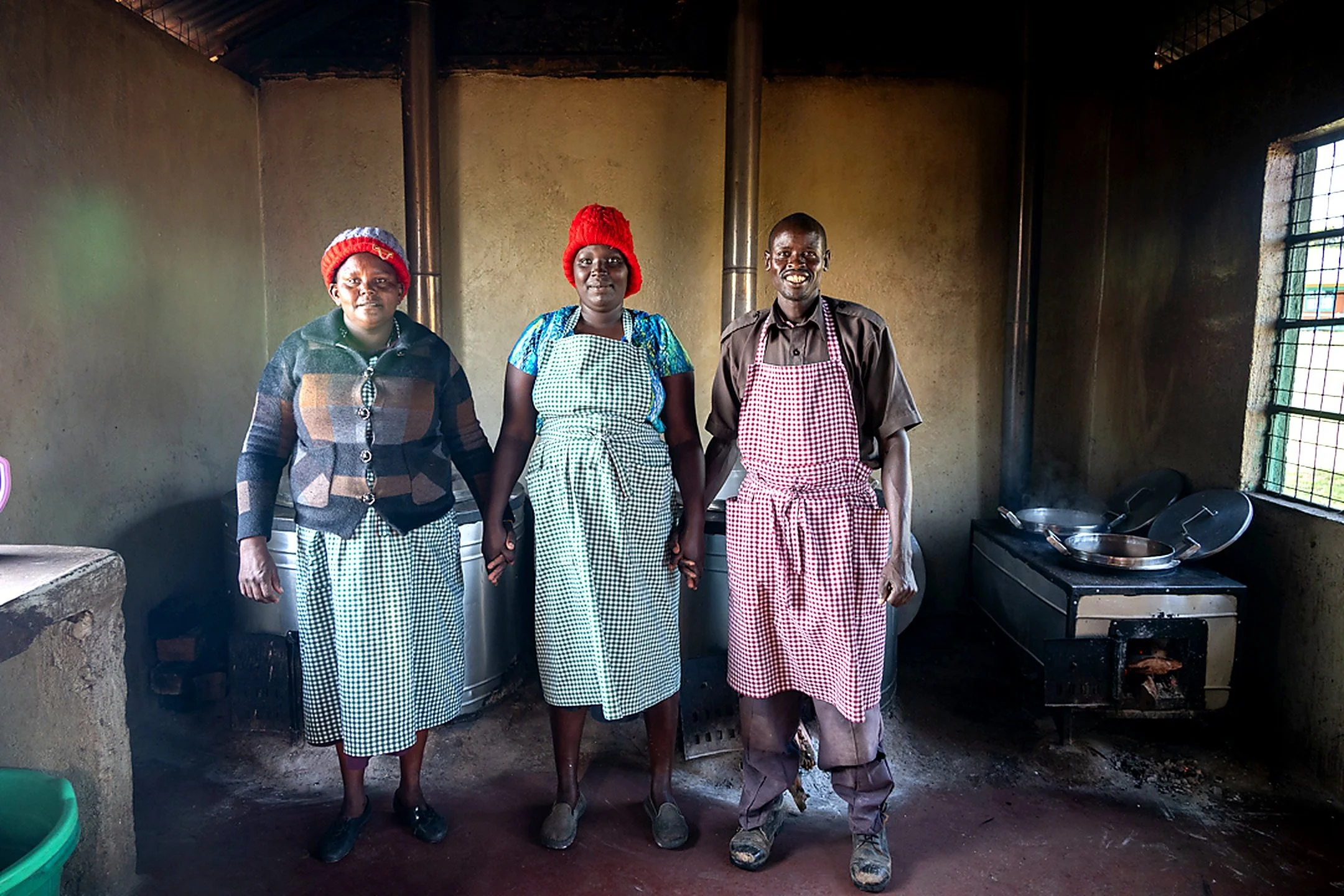 Four people stand in a kitchen, three women and one man, all wearing aprons and the women wearing red knit hats. The kitchen has a stove with pots and pans to the side, light streaming through barred windows, and a concrete floor.