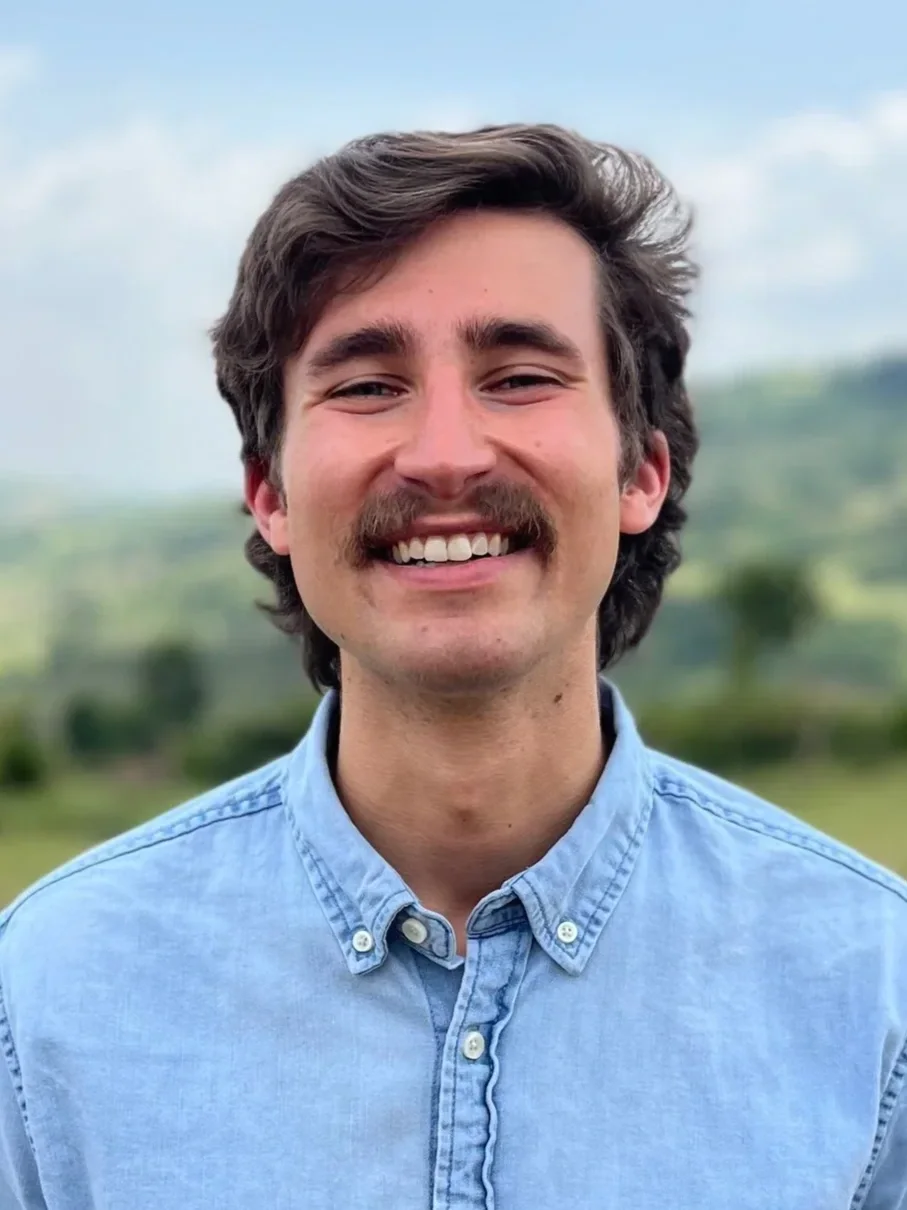 A young man with dark, wavy hair, a mustache, wearing a light blue button-up shirt, smiling outdoors with a blurred green landscape in the background.
