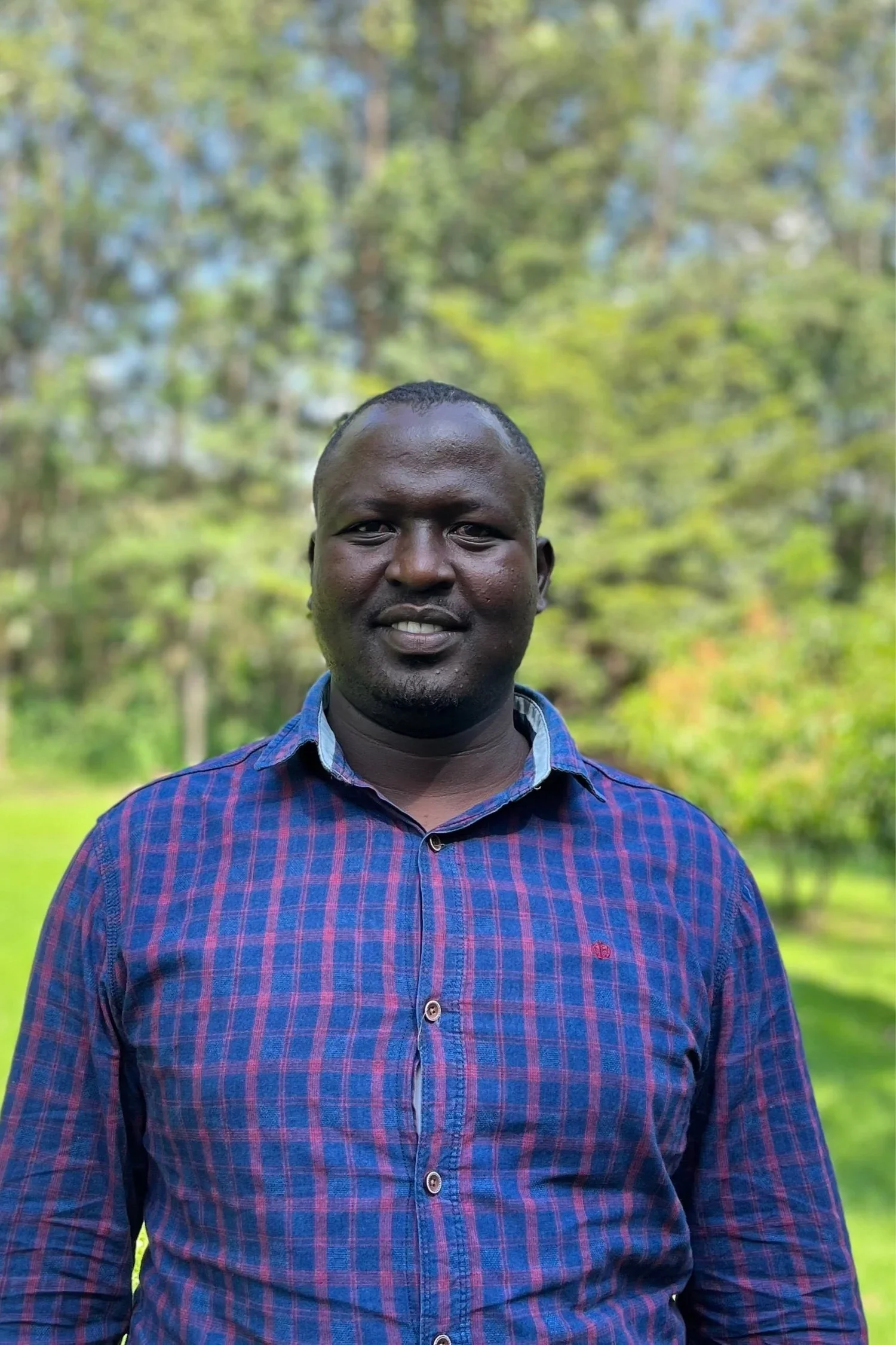 A smiling man with dark skin and short hair wearing a blue and red checkered shirt, standing outdoors with green trees in the background.