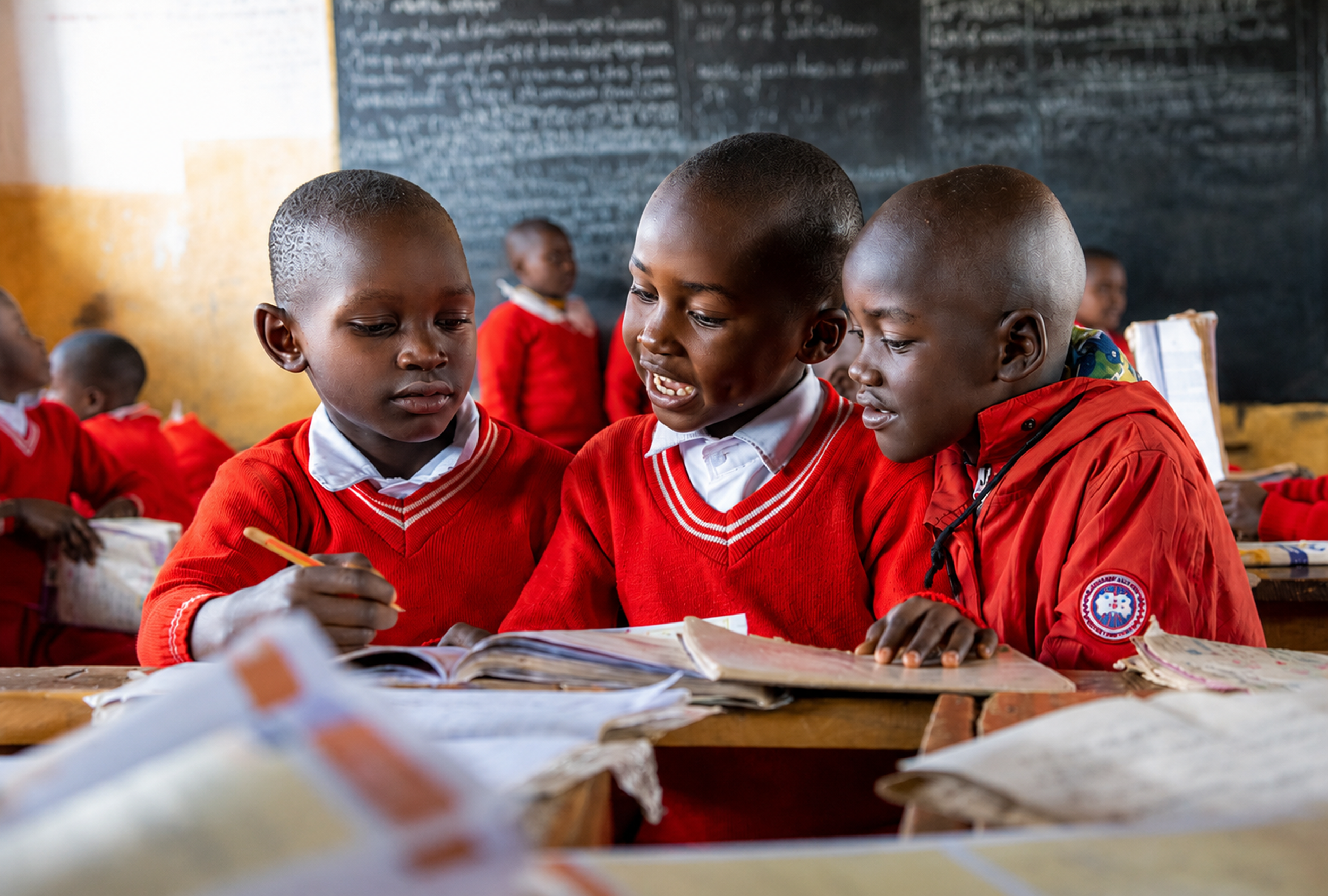 Three school children in red uniforms studying together at a wooden desk in front of a black chalkboard.