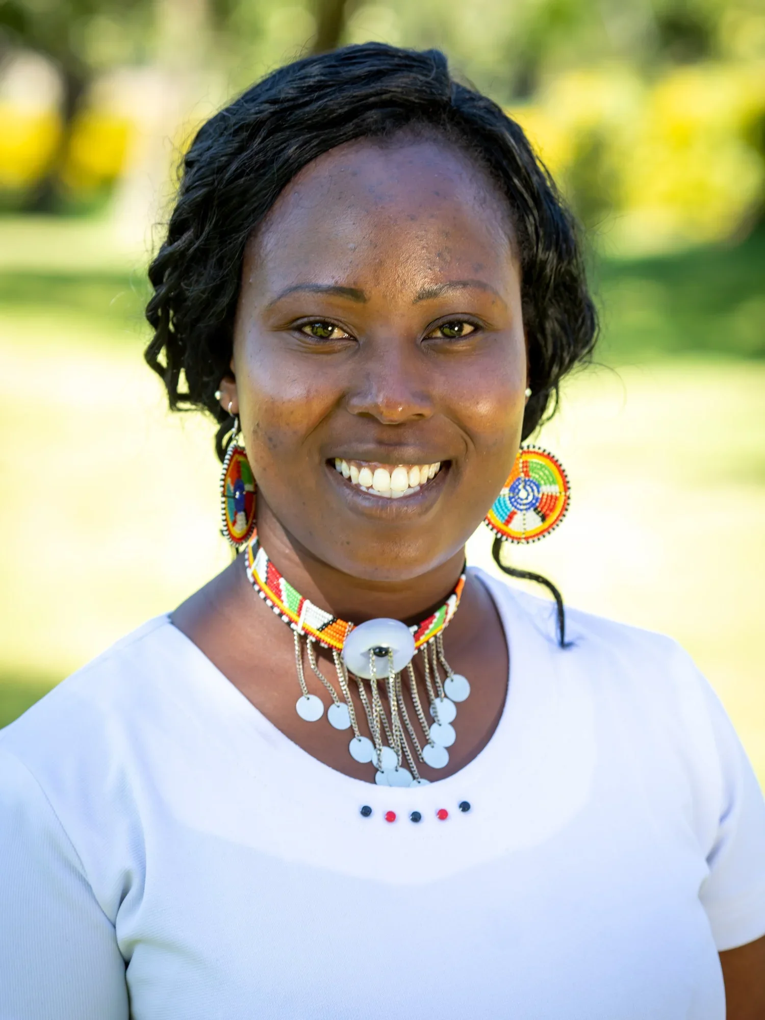 A woman with dark curly hair, smiling outdoors in a park with green trees in the background. She wears colorful beaded jewelry, including large earrings, a necklace, and a decorative choker with white and black accents.