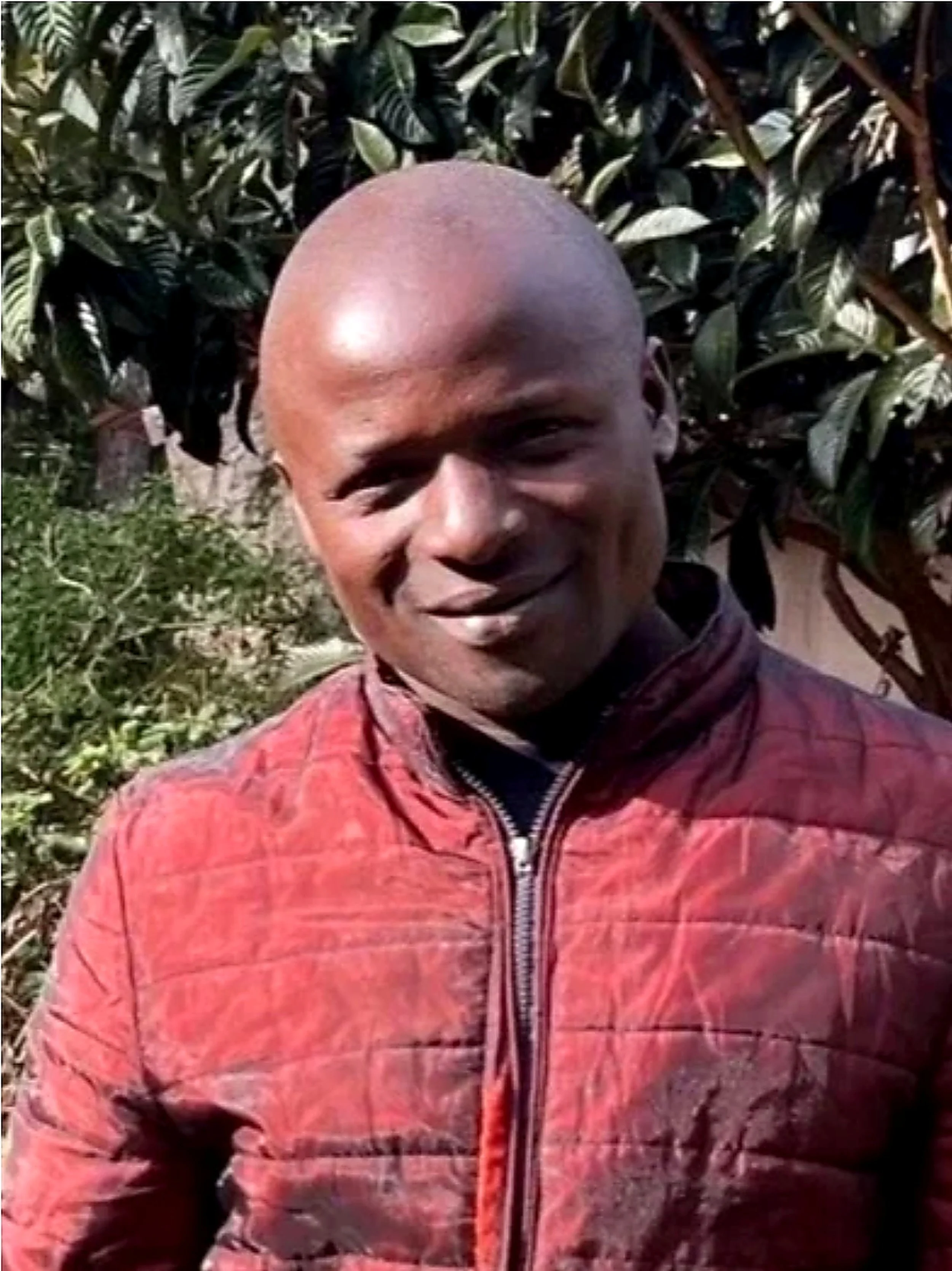 A smiling man with a shaved head wearing a red patterned jacket standing outdoors in front of green foliage.