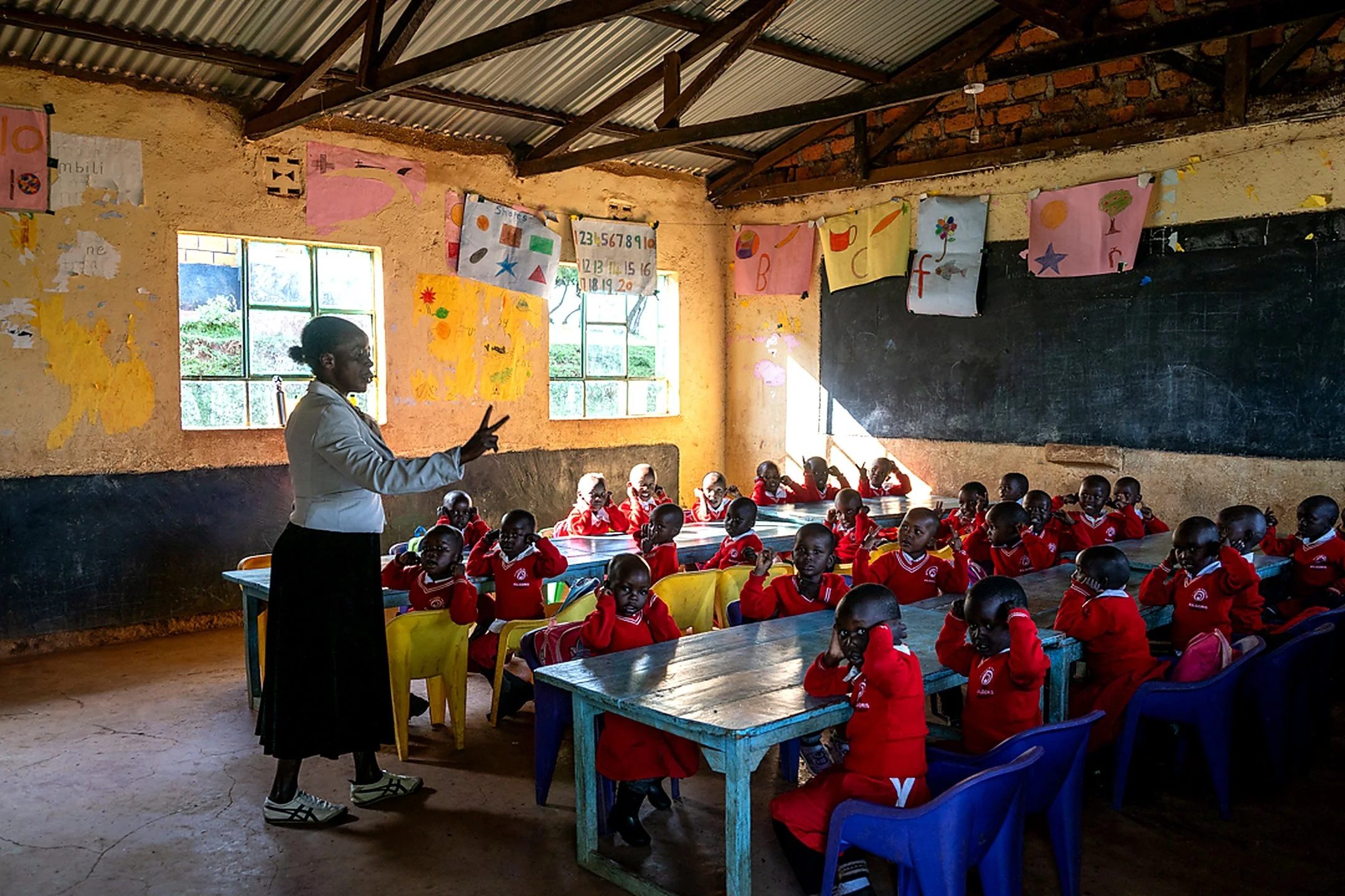A classroom with a teacher standing in front of a blackboard and students wearing red uniforms seated at desks, in a room with corrugated metal roof and windows, decorated with colorful artwork and educational charts.