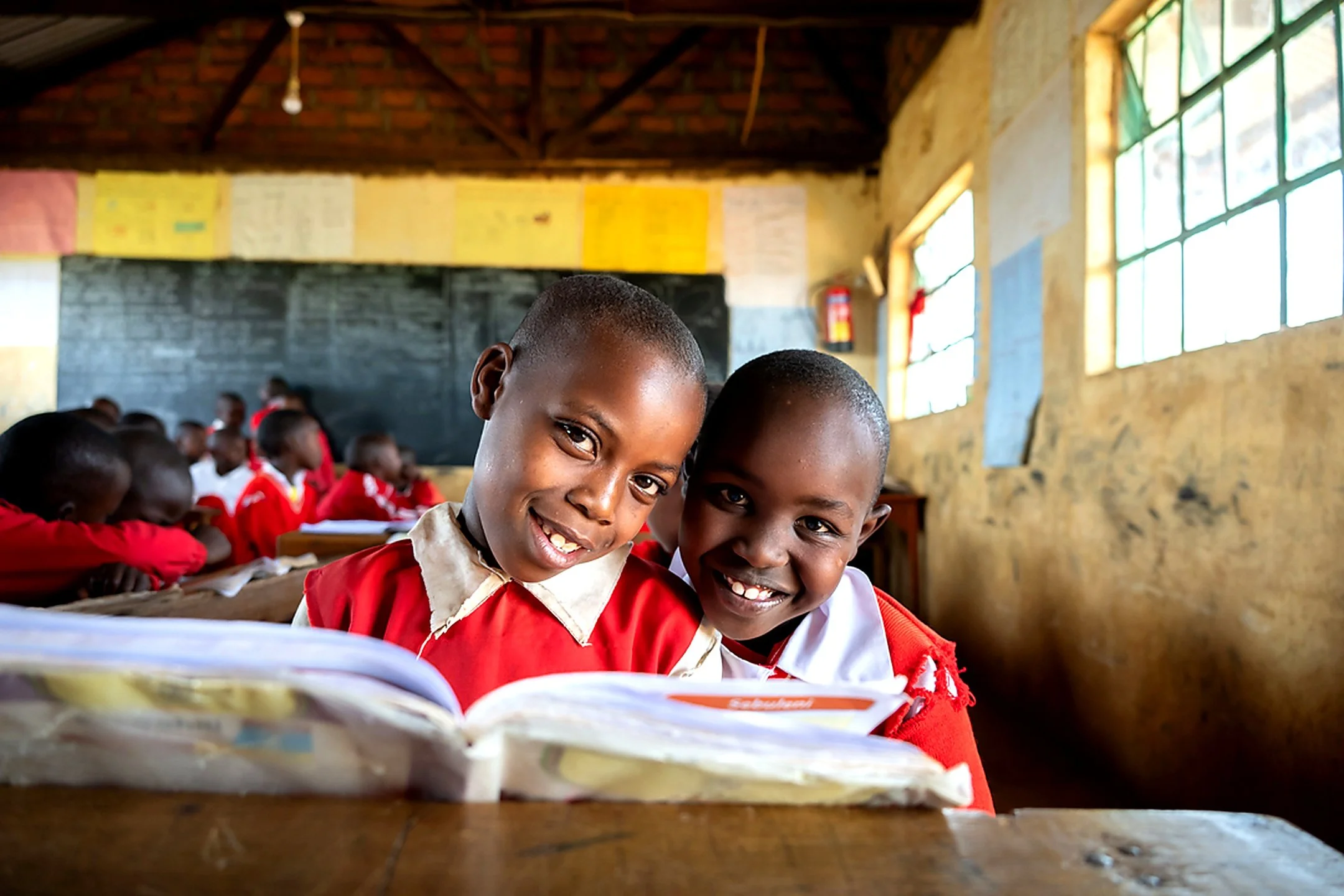 Smiling school children in red and white uniforms sitting at desks in a classroom with brick and concrete walls and large windows.
