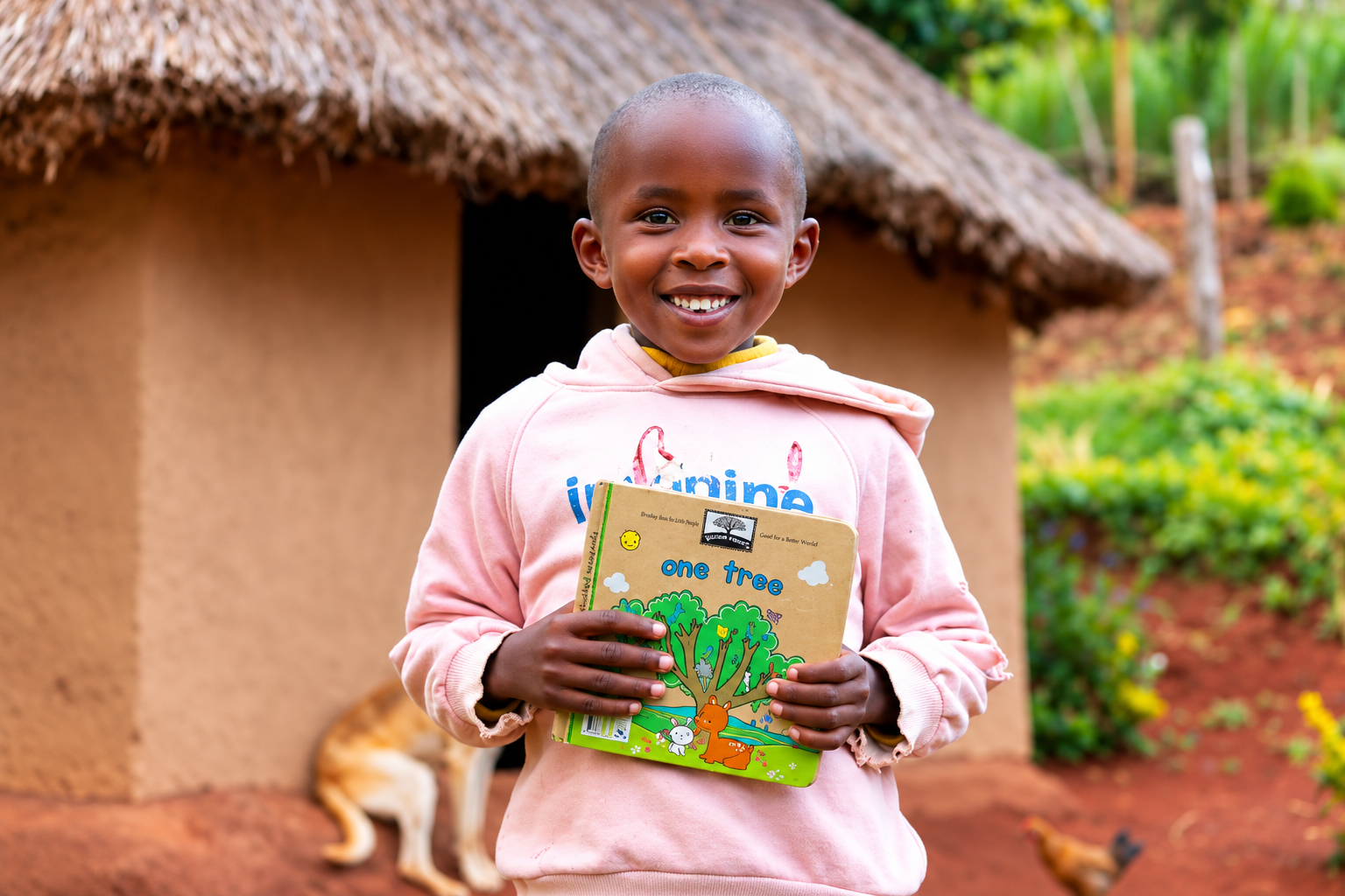 Smiling child holding a colorful “One Tree” children’s book in front of a thatched-roof home