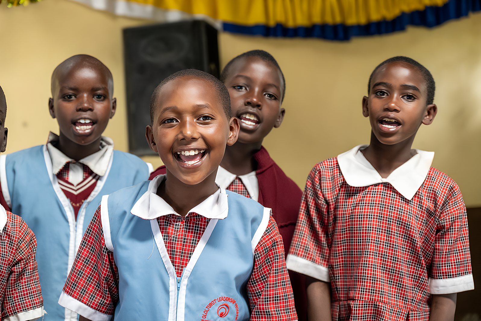Smiling children in school uniforms standing together indoors, with a yellow wall and a speaker in the background.