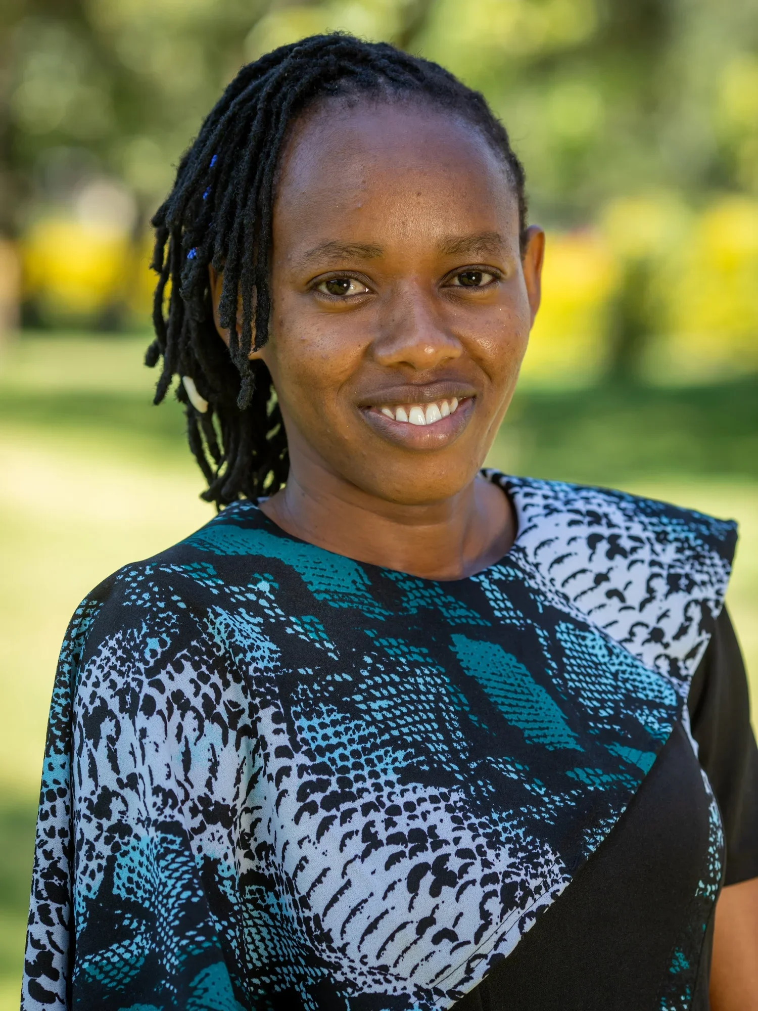 A woman with short dreadlocks, wearing a patterned black, white, and blue top, is smiling outdoors with a blurred green background.
