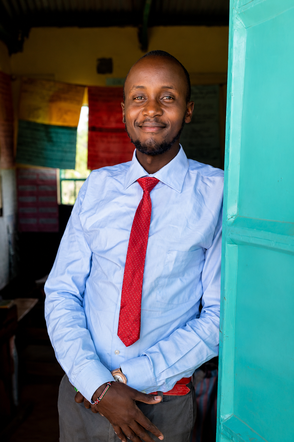 A man wearing a light blue dress shirt, red tie with small white dots, and gray pants stands smiling in a doorway, with colorful fabric banners in the background.