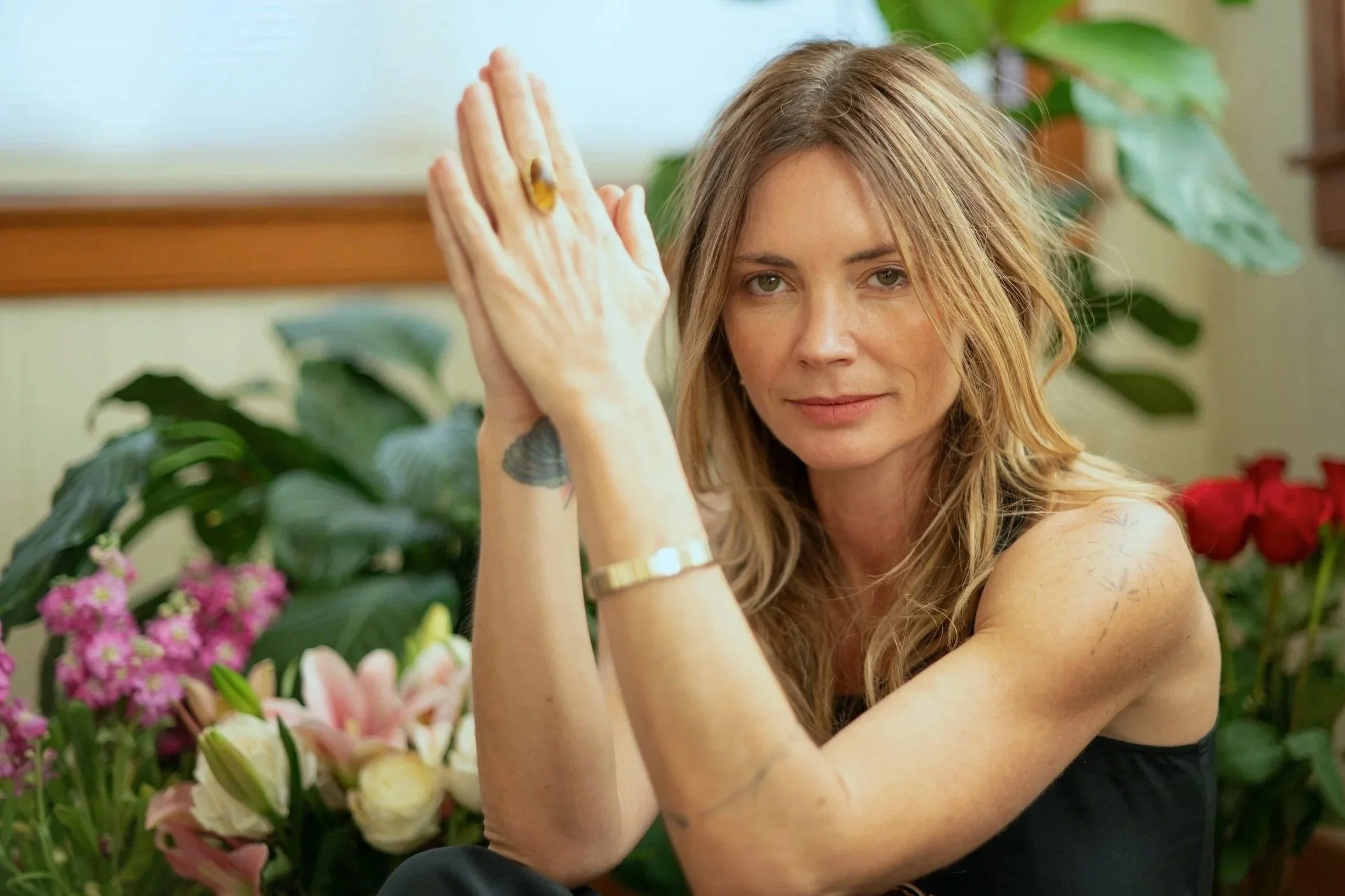 A woman with blonde hair and tattoos sitting in front of colorful flowers, looking at the camera with a relaxed expression.