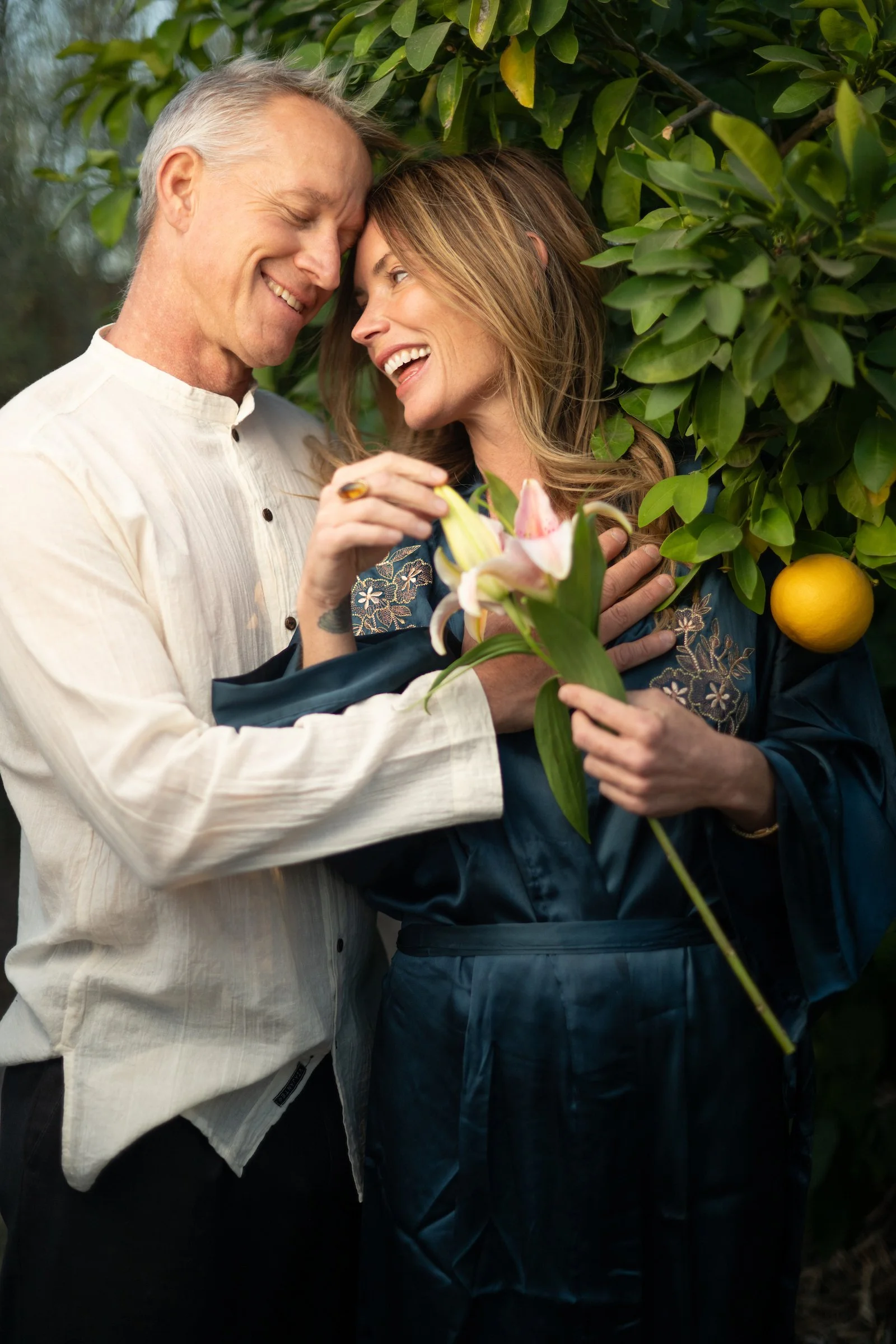 A happy older man and a woman sharing a joyful moment outdoors, surrounded by green foliage. The man has gray hair and is dressed in a light-colored shirt, while the woman has long, light brown hair and is holding a bouquet of lilies. They are close together, smiling and touching foreheads.