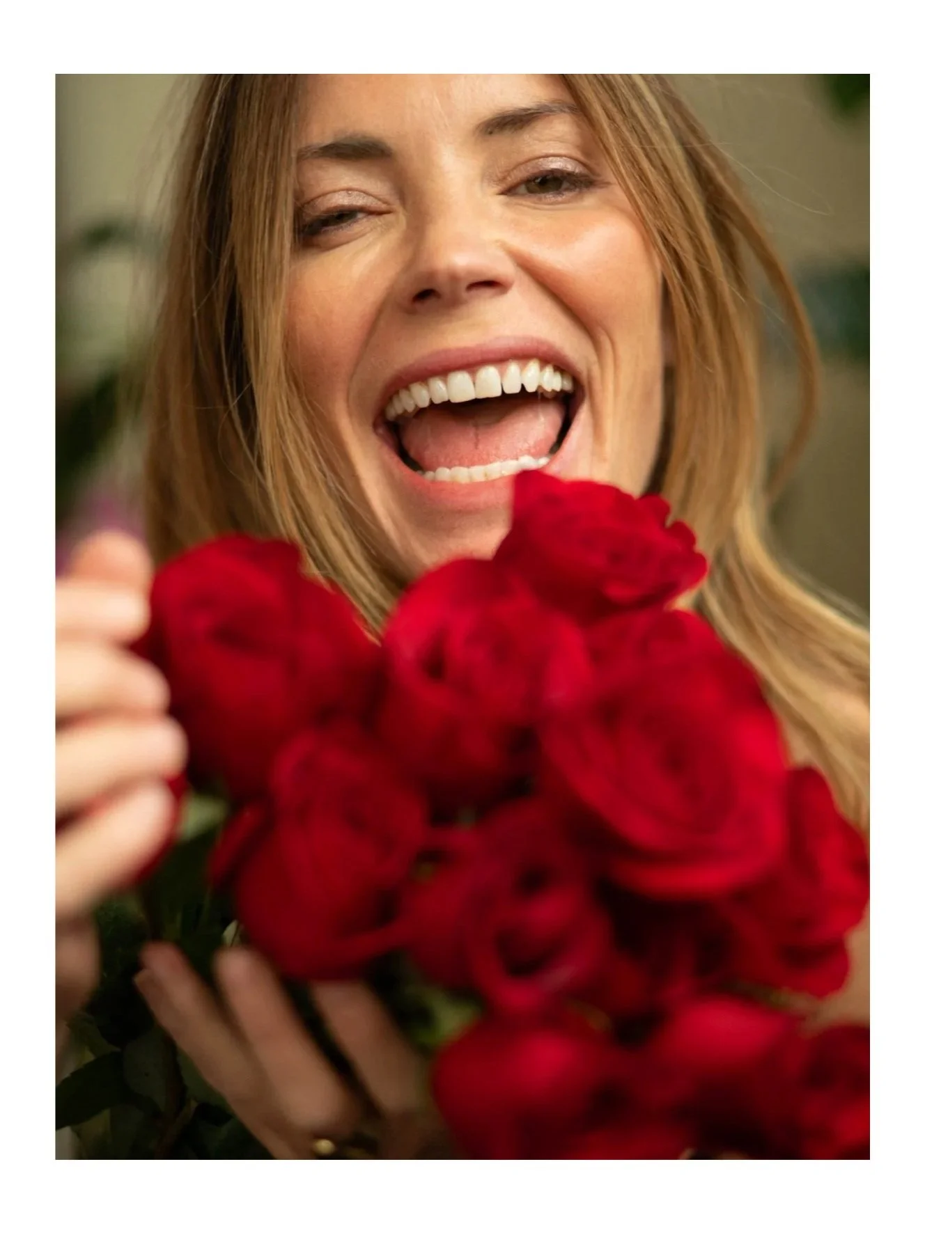 A woman with long red hair holding a bouquet of red roses, smiling and looking joyful.