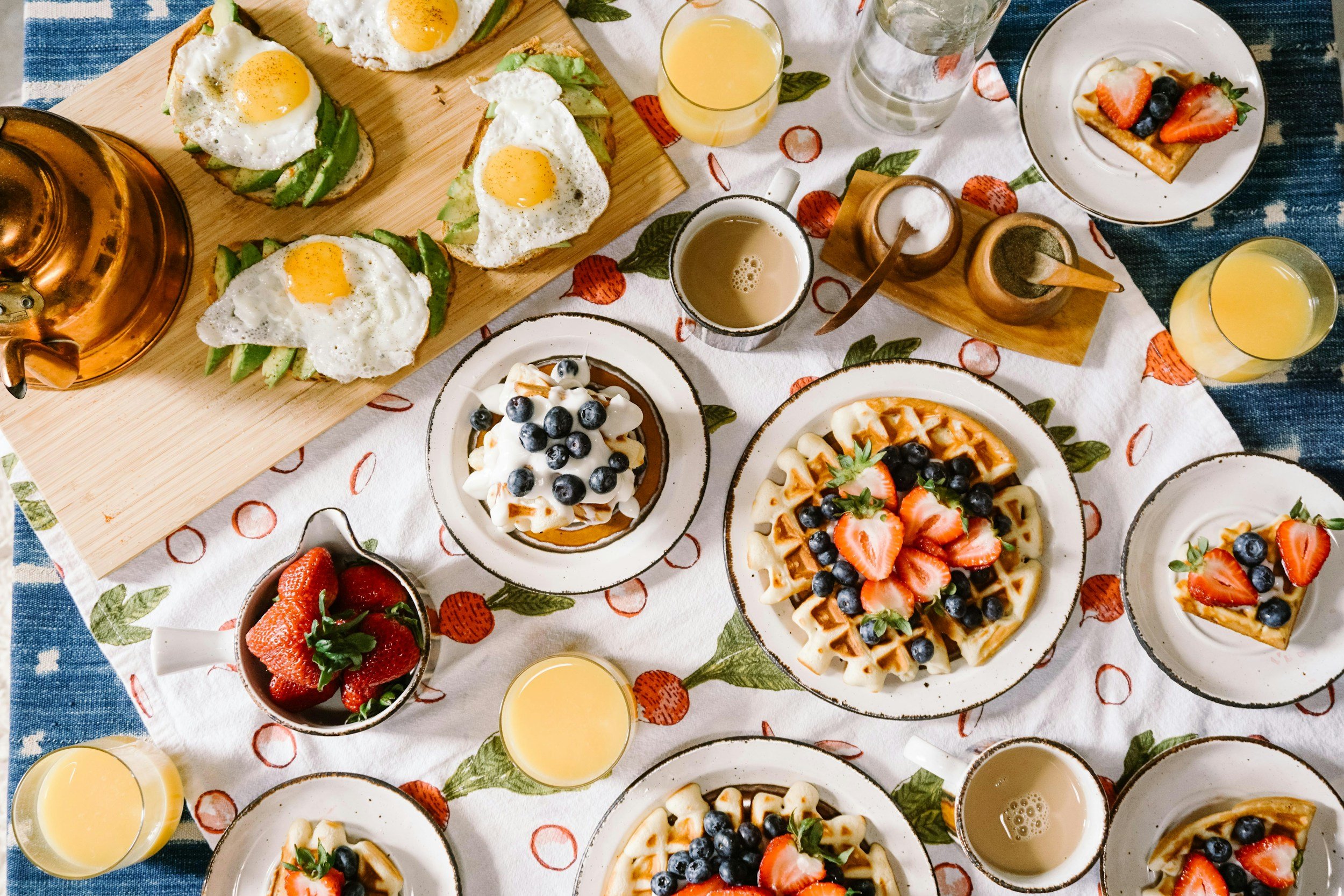 A breakfast spread including waffles topped with blueberries and strawberries, avocado toast with fried eggs, small bowls of strawberries, and glasses of orange juice on a table with a patterned tablecloth.